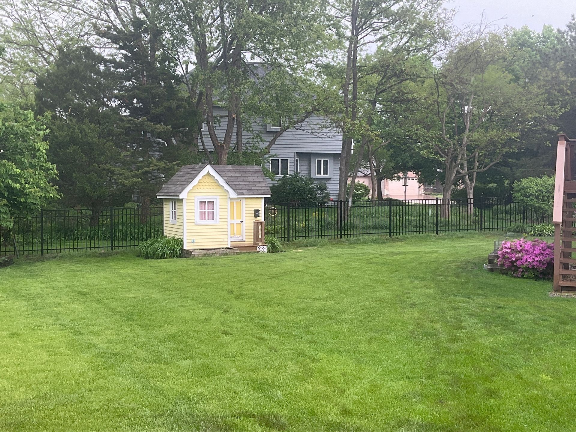 Yellow playhouse in a fenced backyard with green grass and trees, a gray house in the background.