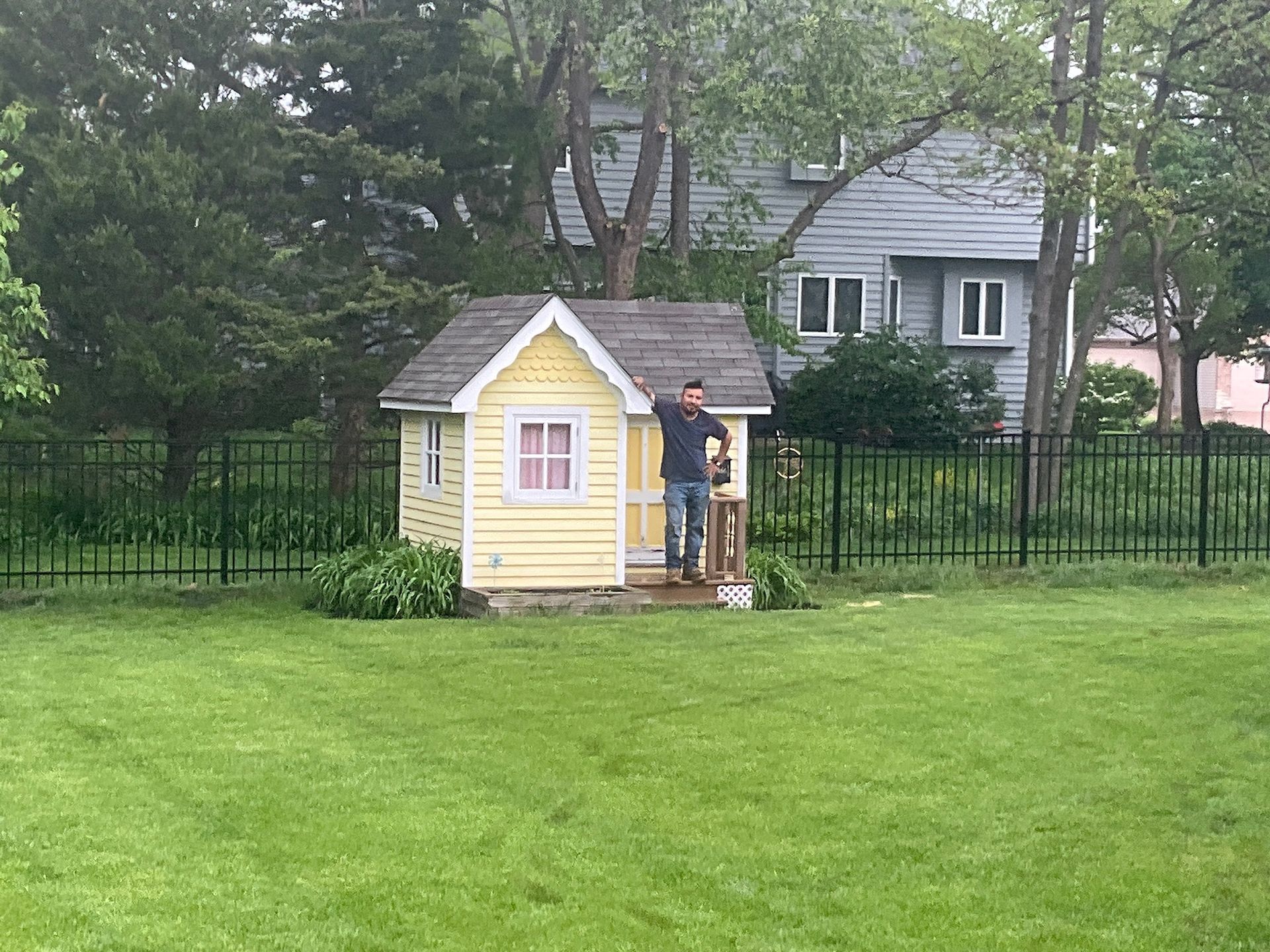 Man standing in front of yellow playhouse in yard with a house and trees in the background.