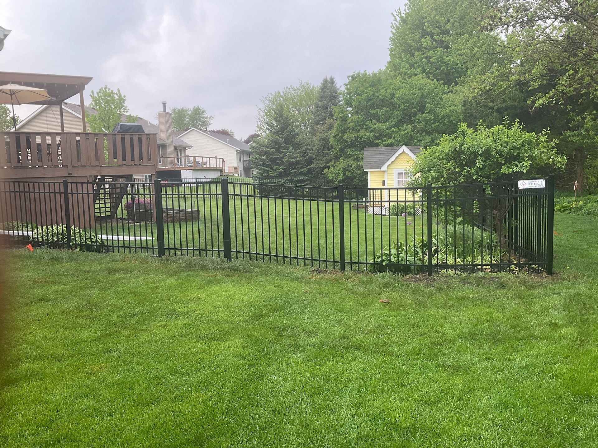 Black metal fence in a backyard with a playset and a small yellow building. Green grass and trees are in the background.