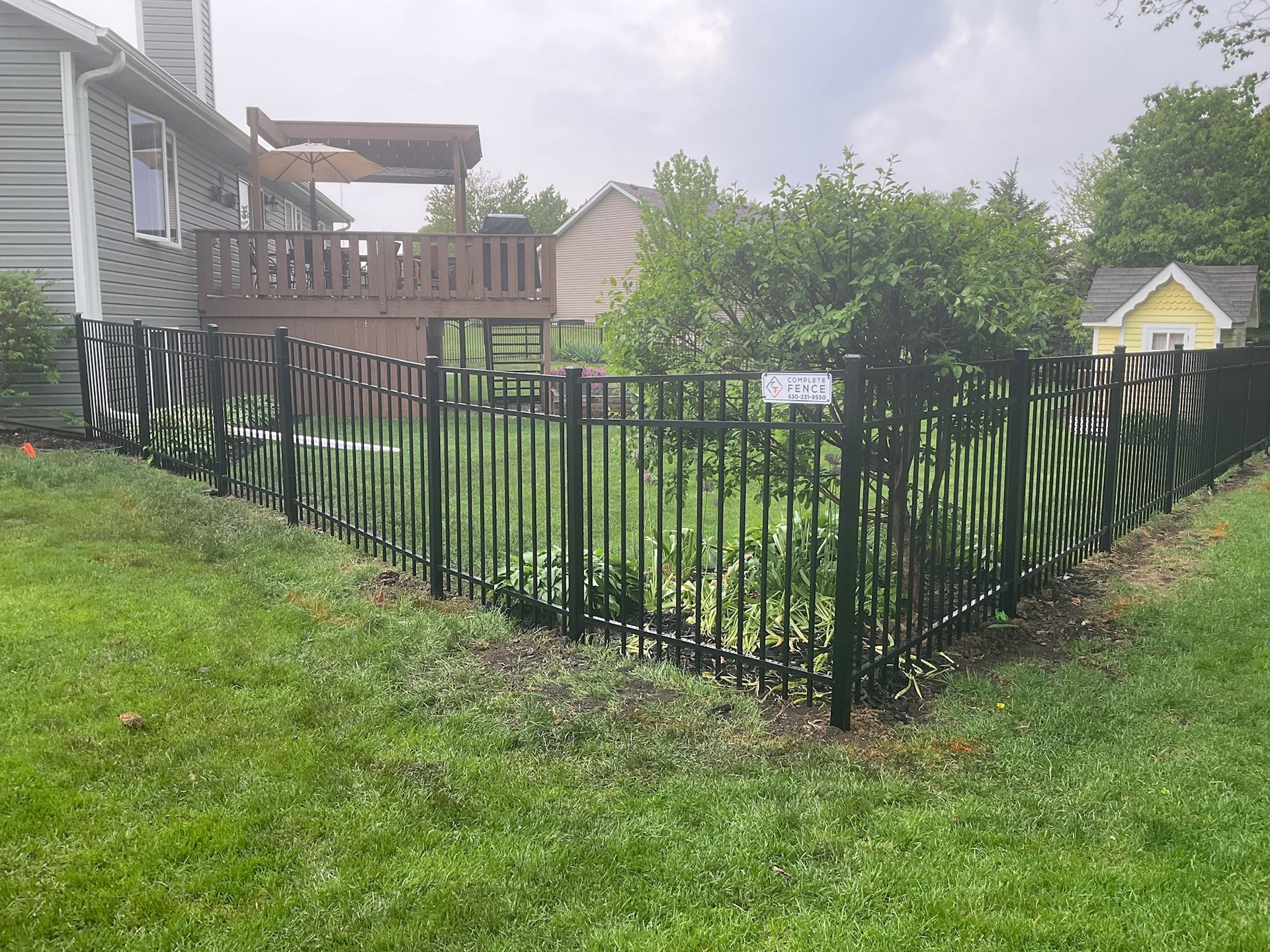 Black metal fence surrounds a grassy yard, partially encircling a deck attached to a house. Cloudy sky overhead.