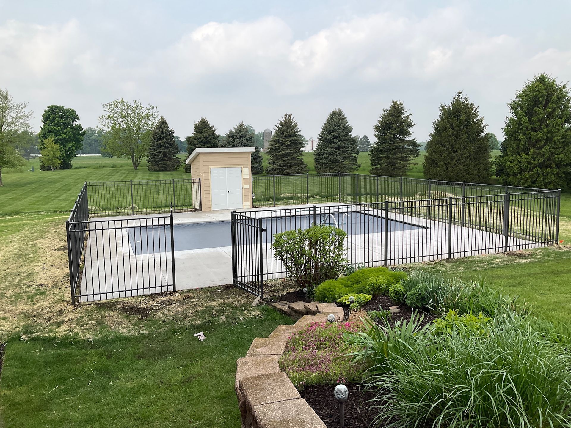 A fenced-in rectangular swimming pool with a small beige shed, surrounded by grass and trees under a cloudy sky.