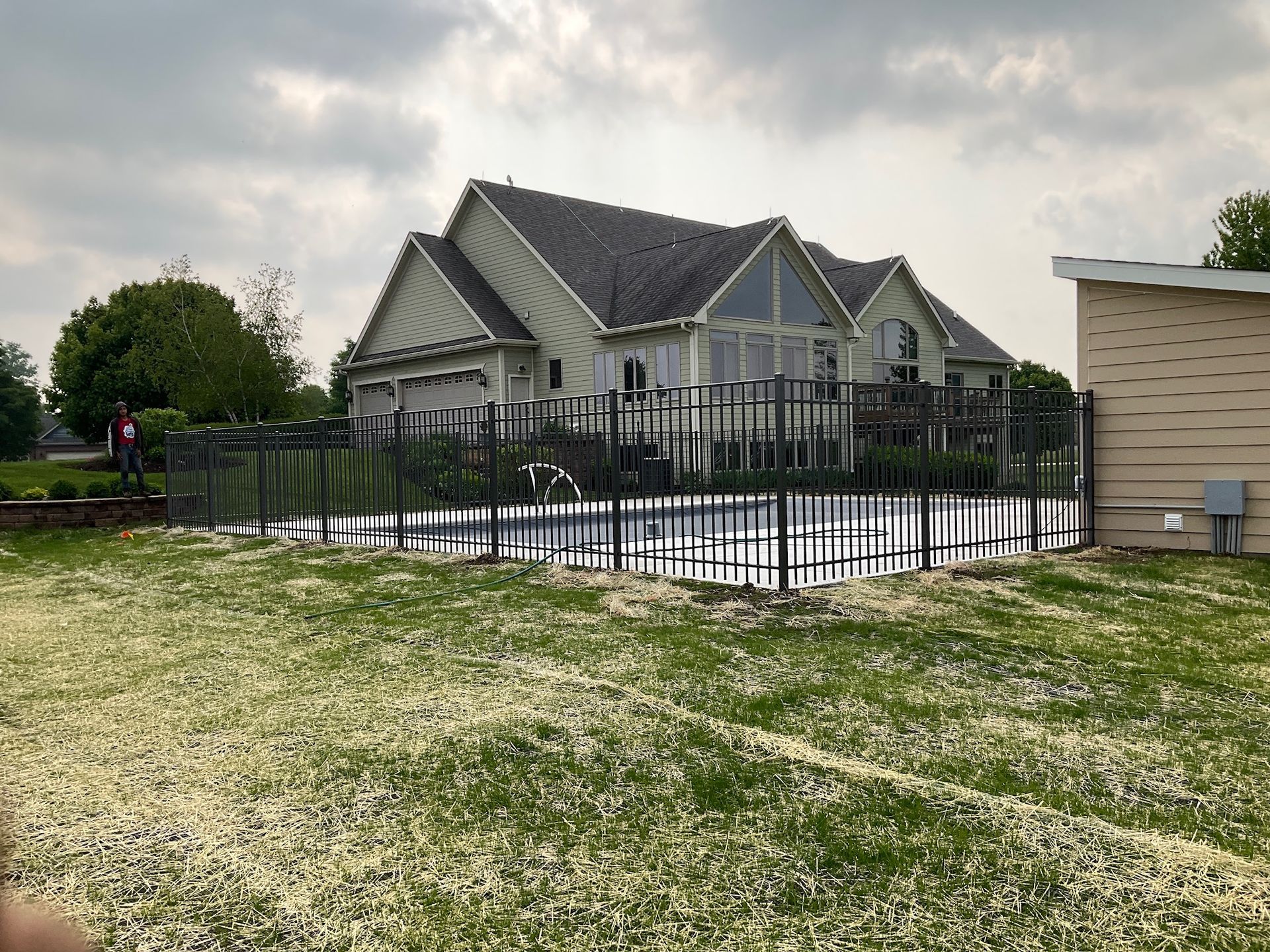 Backyard pool enclosed by black metal fence; house in background. Green grass covered in white debris. Overcast sky.