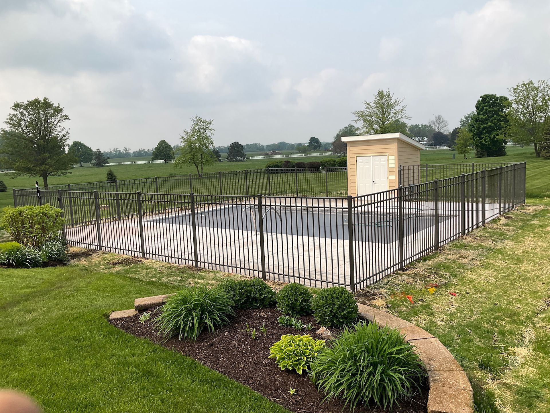 A rectangular pool enclosed by a black metal fence, a small beige shed, and a well-manicured lawn.