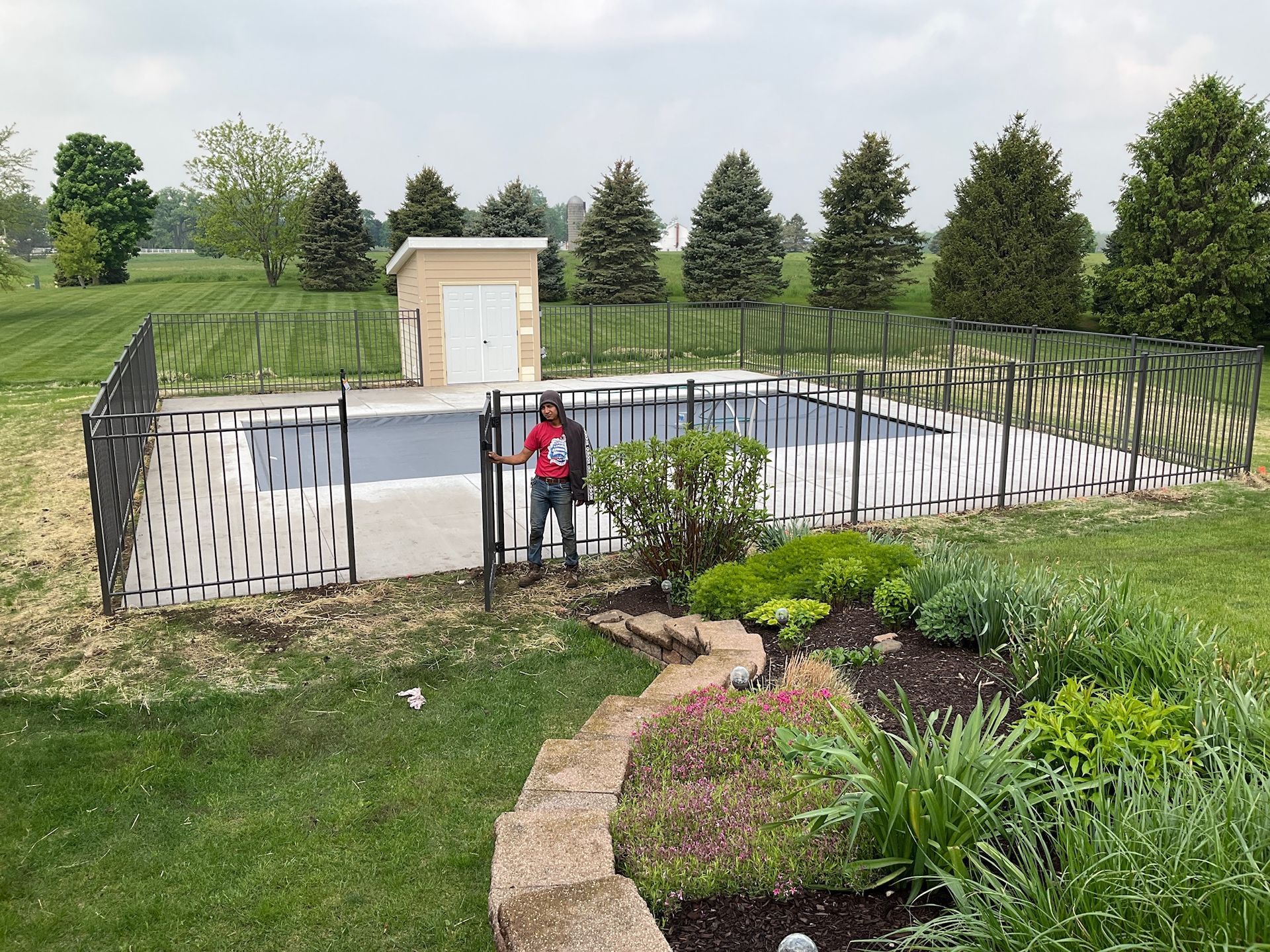 Person stands by a pool with a decorative black fence, small building, and landscaping in yard.