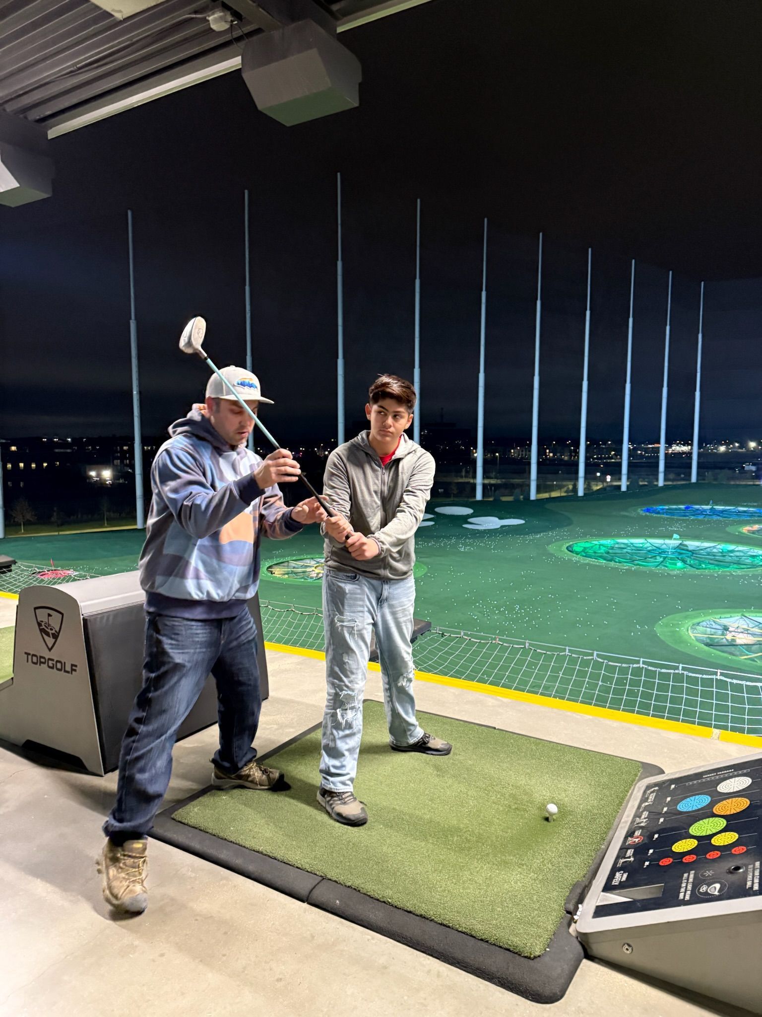 Man coaching a person at a Topgolf, outdoors at night. The man holds a golf club while the person practices the swing.