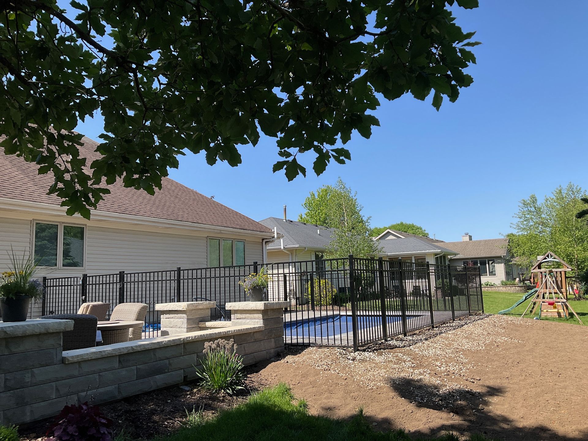 Backyard with a pool, patio, house, and black fence on a sunny day.