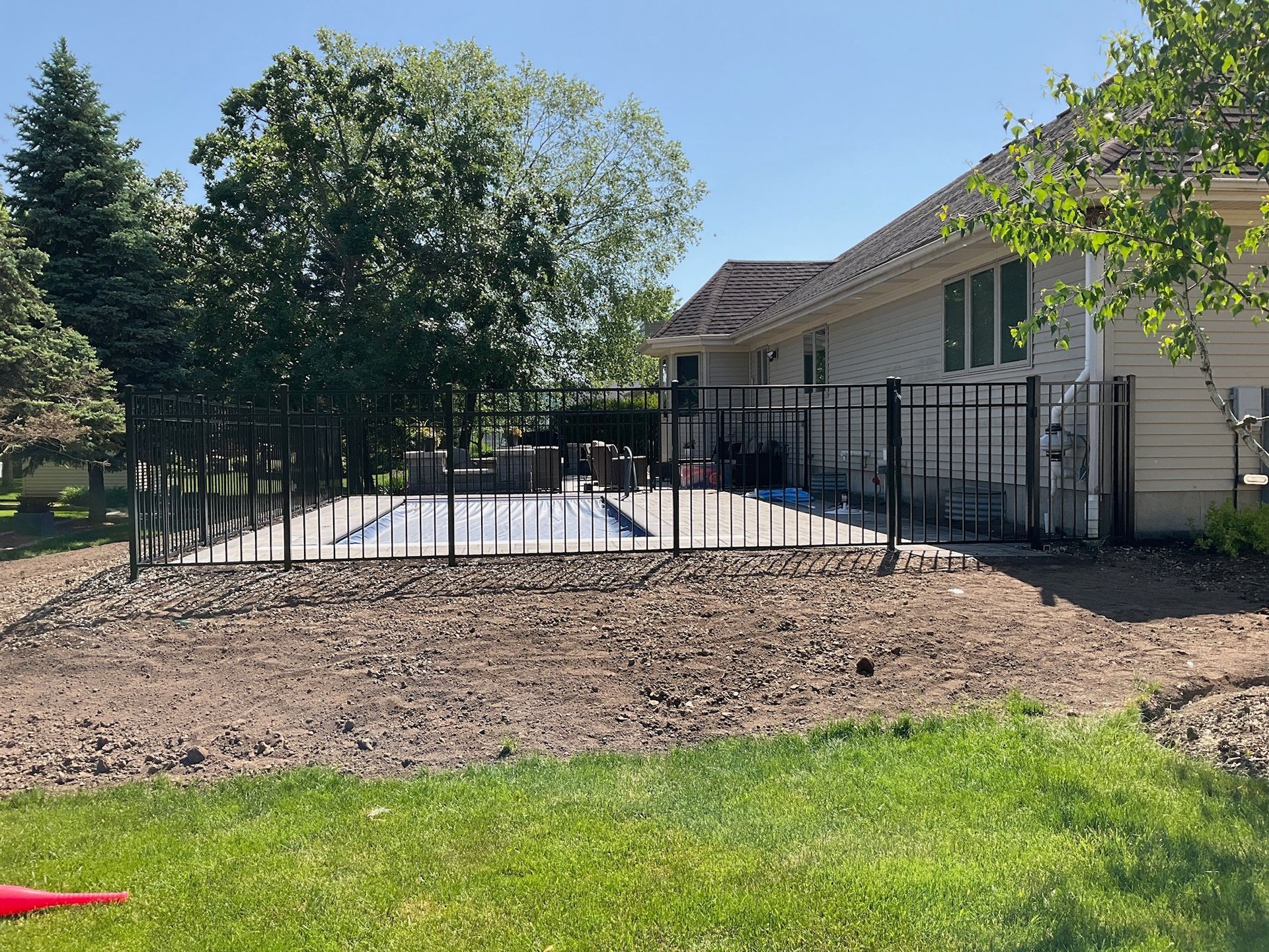 Black fenced-in pool area next to a house with a brown roof and a dirt/grass yard.
