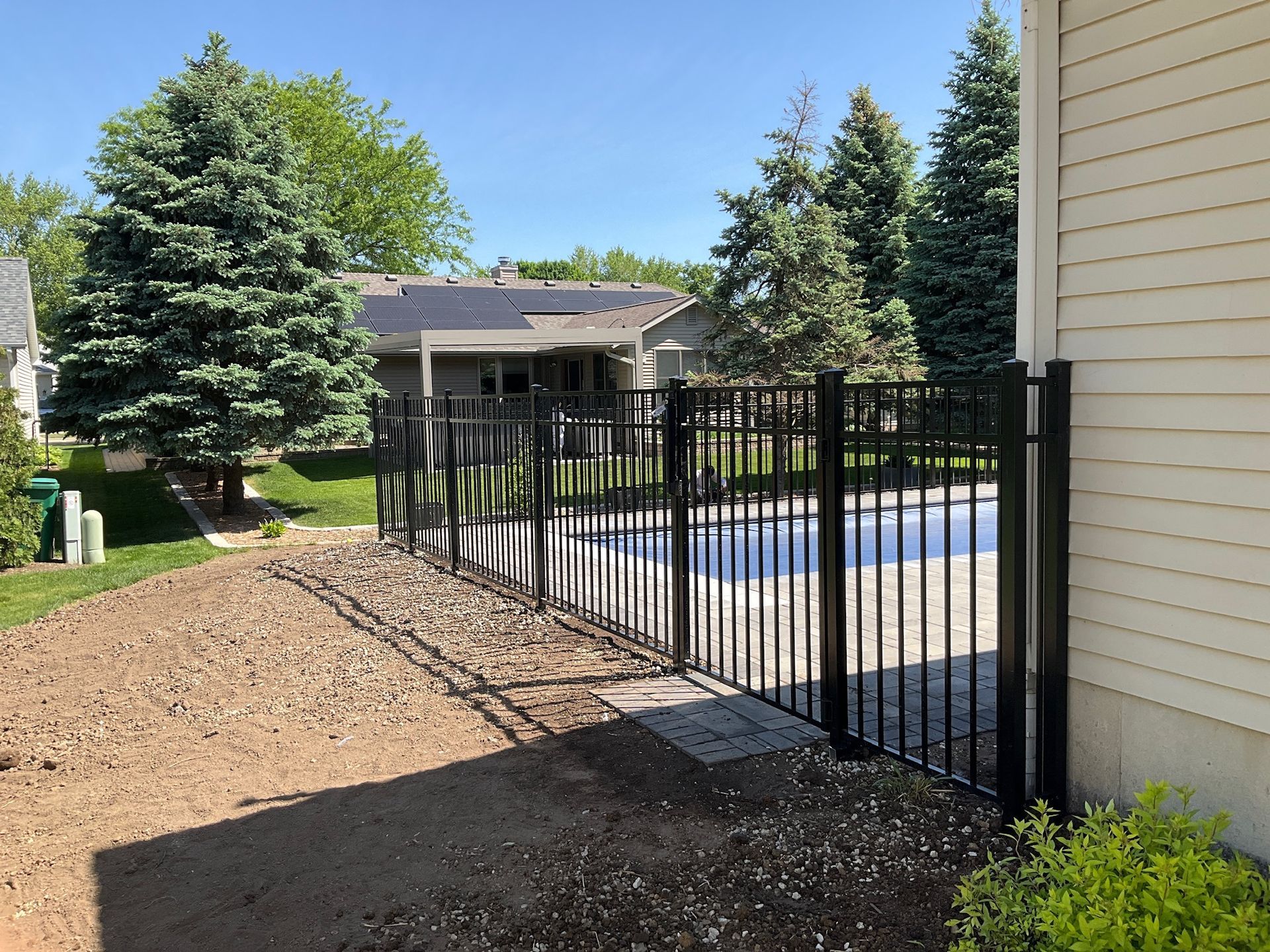 Black metal fence encloses a pool area next to a house with gravel, trees, and blue sky.