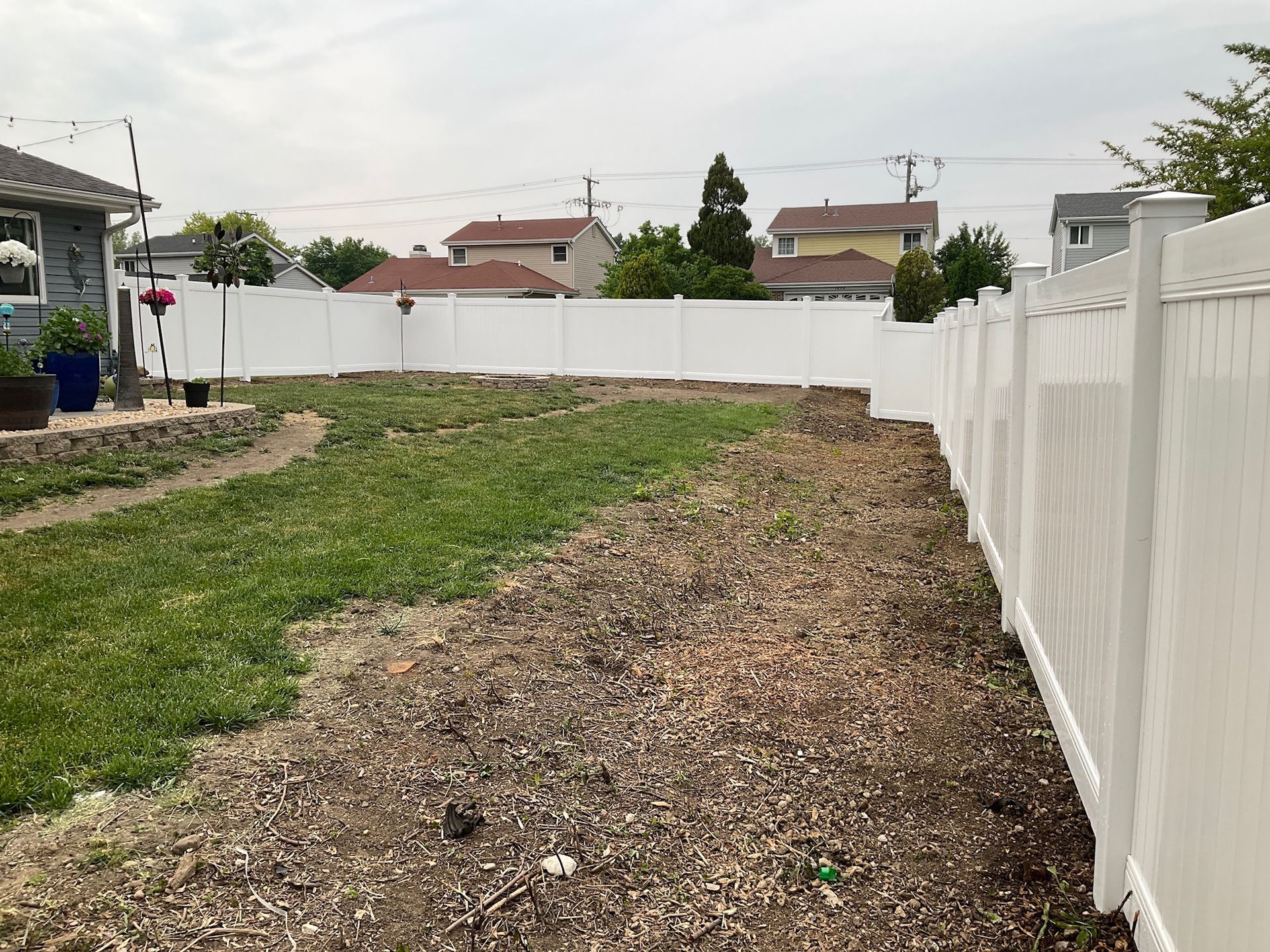Backyard with white vinyl fence, green grass, and a brown mulch area. Houses and sky in background.
