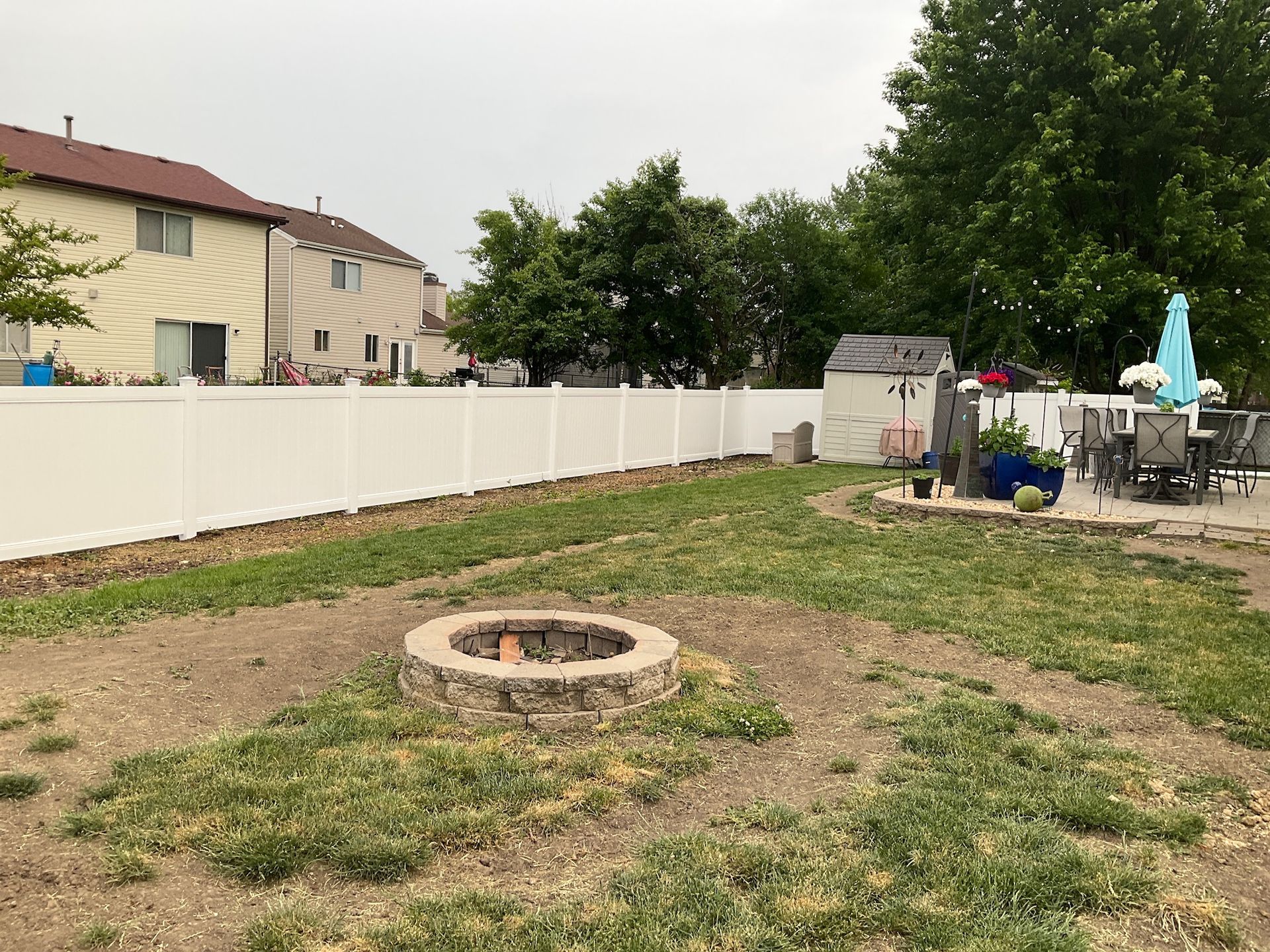 Backyard with white fence, fire pit, shed, patio furniture, and houses. Overcast sky.