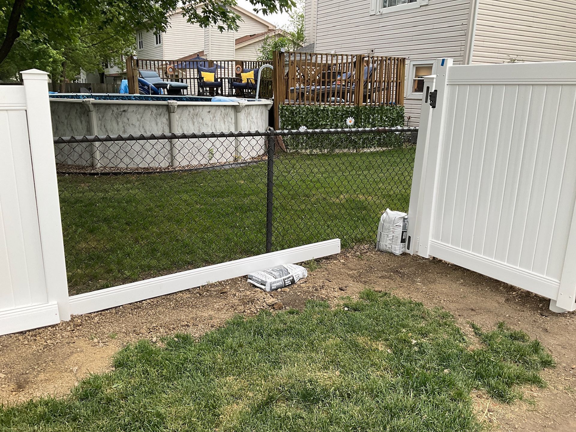 White vinyl fence gate open, leading to a backyard with pool and chain link fence.
