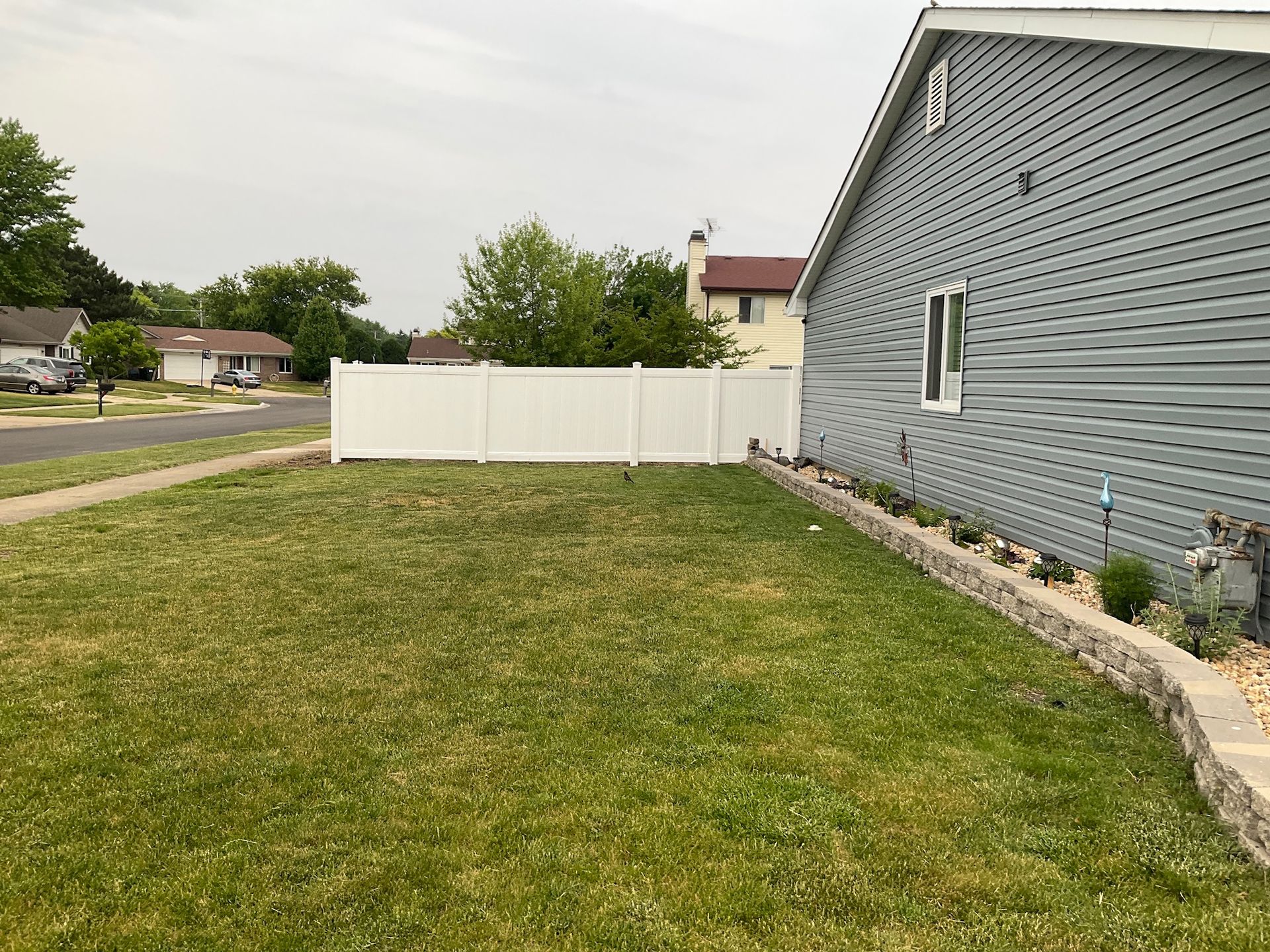 Lawn with a white fence alongside a gray house. Houses in the distance and a retaining wall with plants.