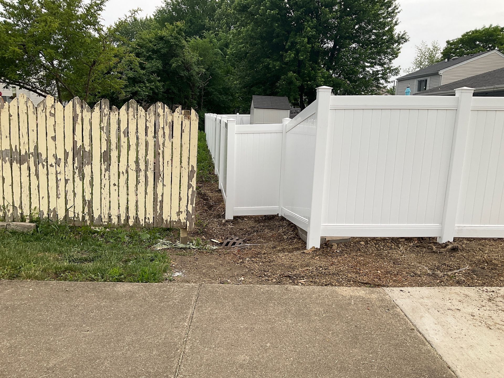 Old peeling yellow fence meets new white vinyl fence and gate; side-by-side on a sidewalk.