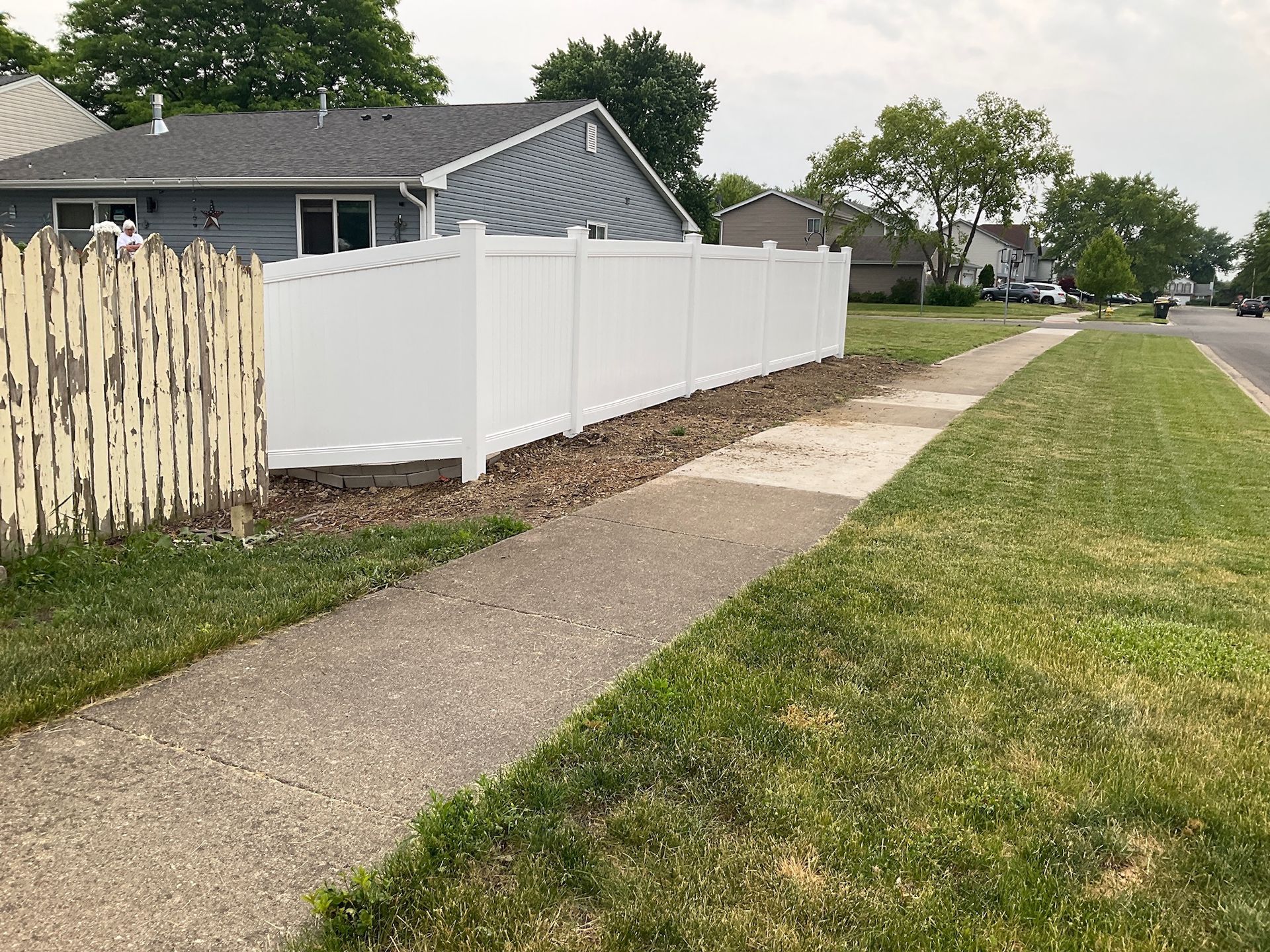 White fence beside a sidewalk and grass, with a weathered wooden fence on the left.