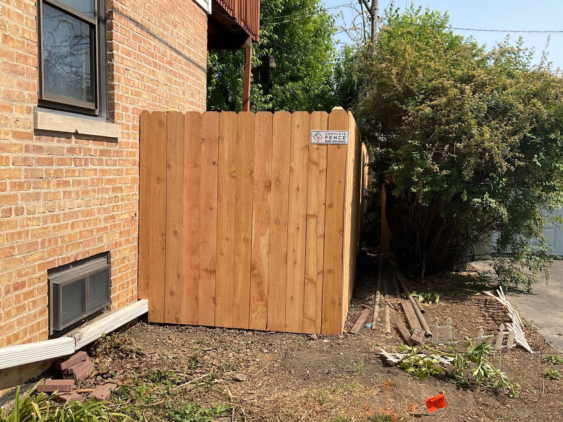 Wooden fence against a brick building, concealing an area with a bush and metal debris.