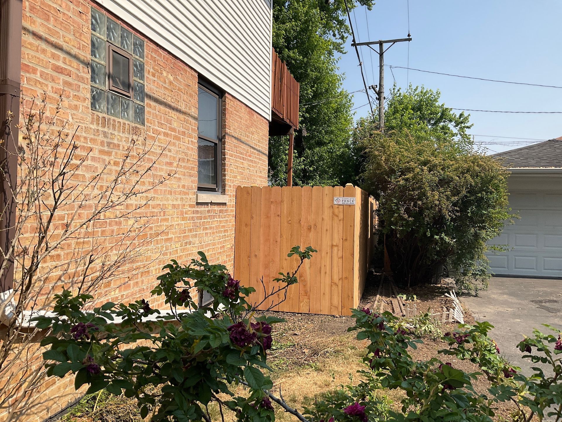 Wooden fence next to brick building, with bushes and garage visible. Sunny outdoor setting.