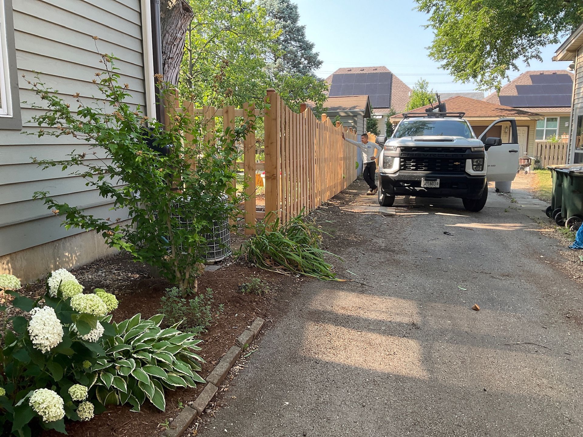 A truck parked in a gravel driveway next to a wooden fence and a house with green foliage.