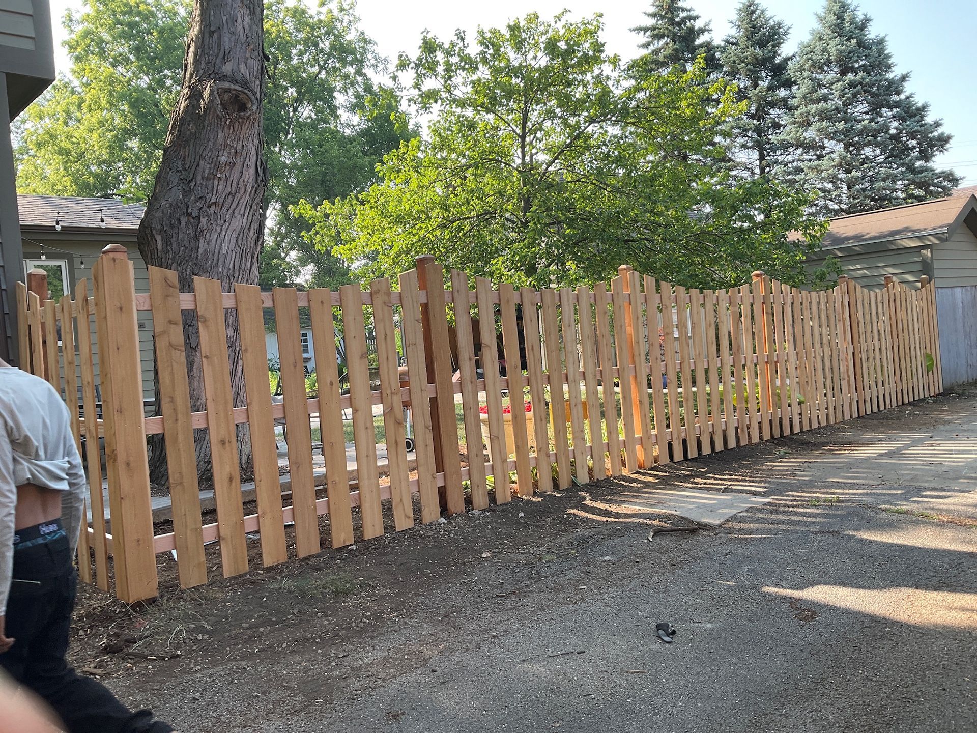 Wooden picket fence in a gravel driveway, with a tree and houses in the background.