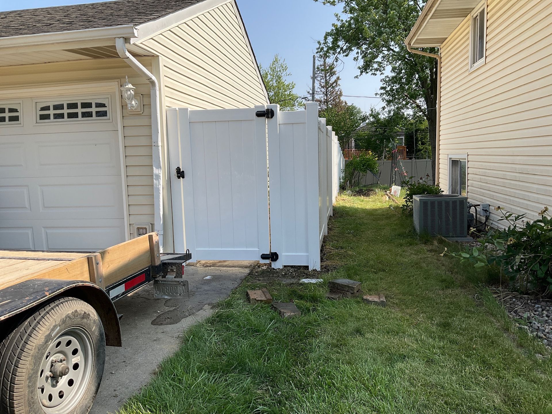 White vinyl fence and gate between two houses. A trailer is in the foreground, with grass in front of the gate.