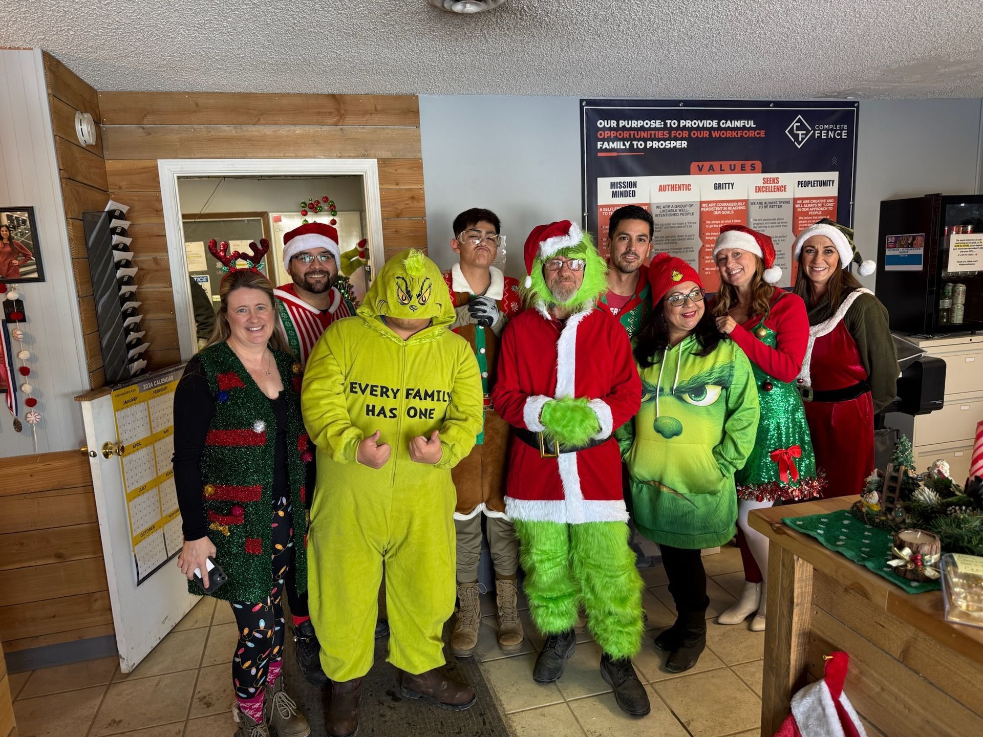 Group of people in Christmas costumes posing indoors, including Grinch, Santa, and elves.