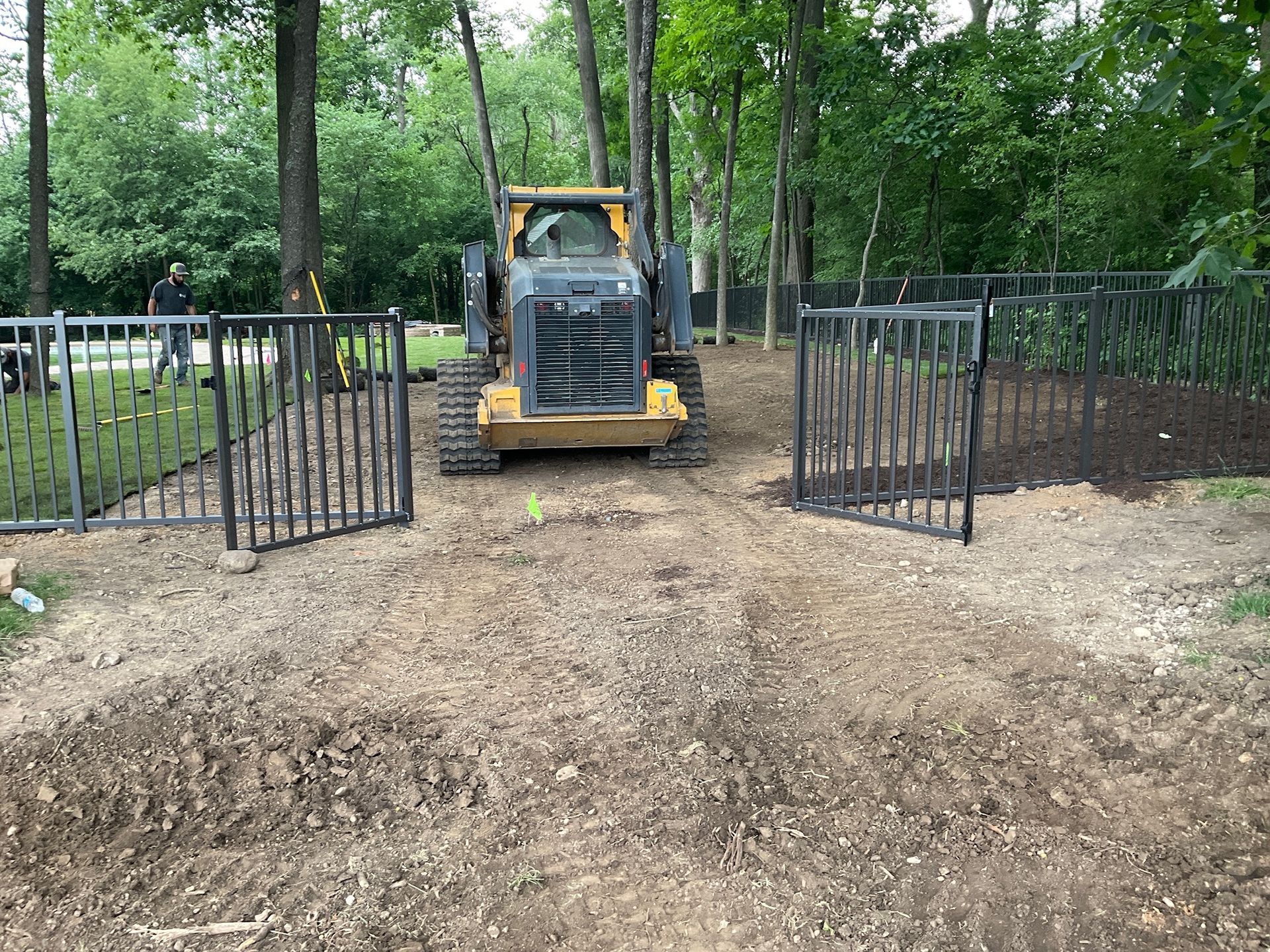 Yellow skid steer in gravel area, open black gate, partially built fence, trees in background.
