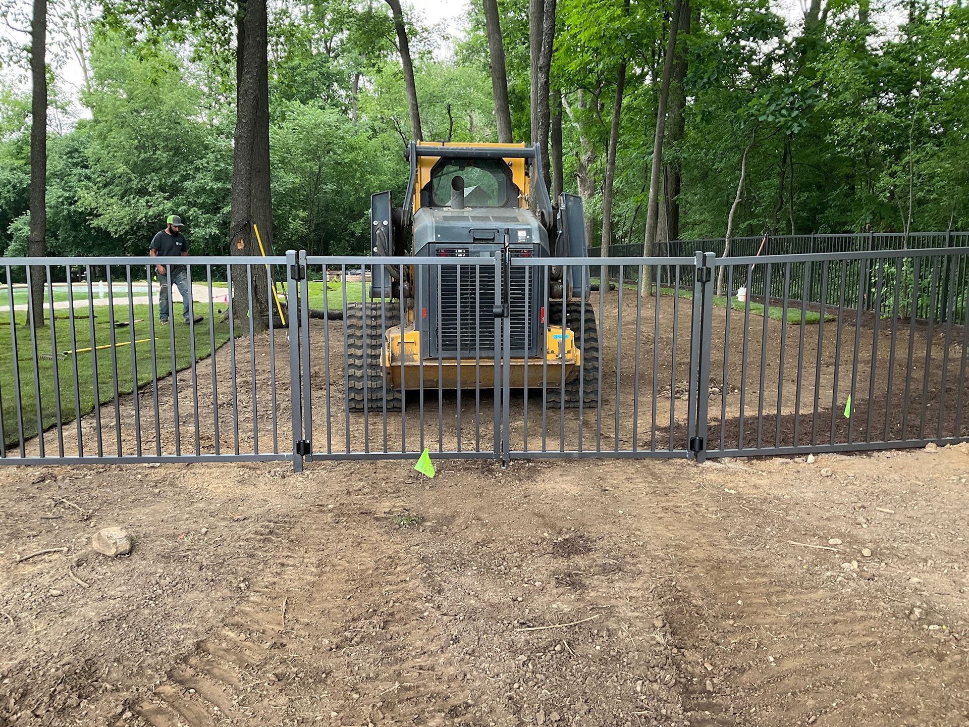 Construction site with a metal fence and yellow front-end loader. A person stands behind the fence in a wooded area.