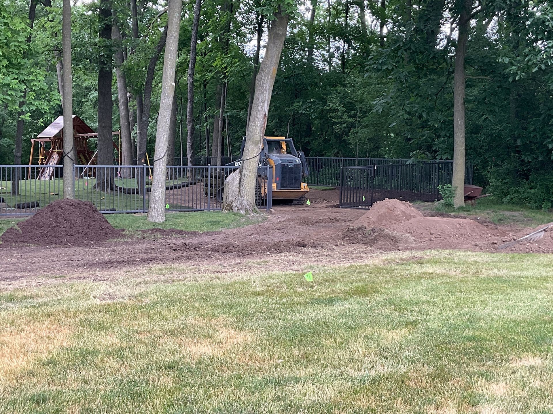 A John Deere tractor is operating near a fence and trees in a yard. Piles of dirt and mulch are present.