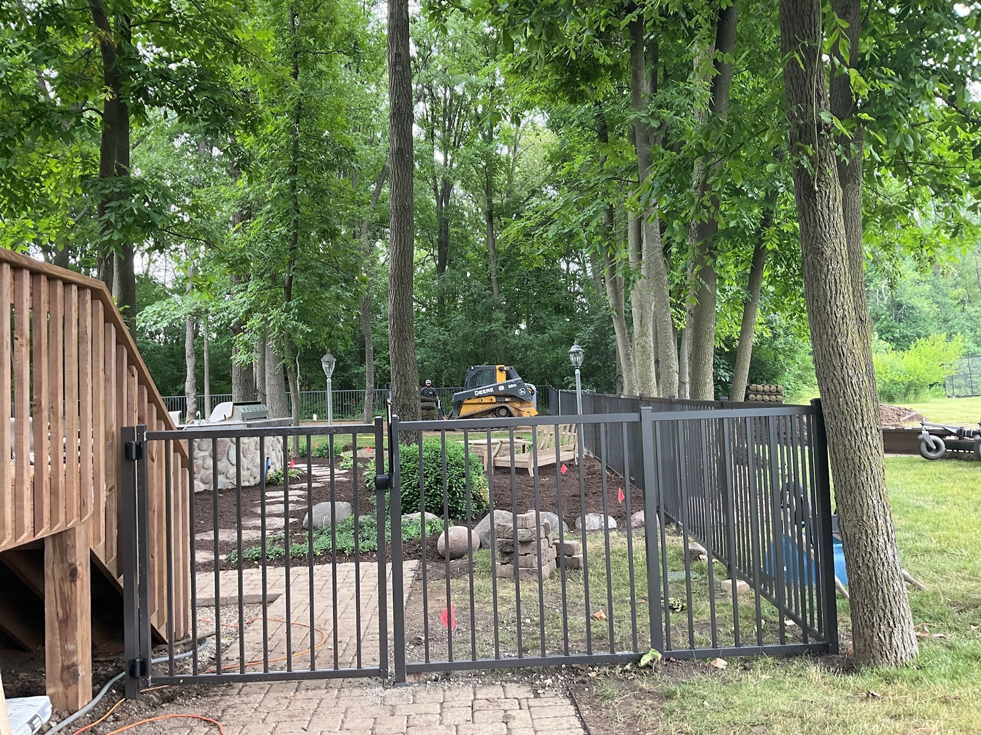 Metal fence encloses a landscaped area with gate, near a wooden deck and trees.