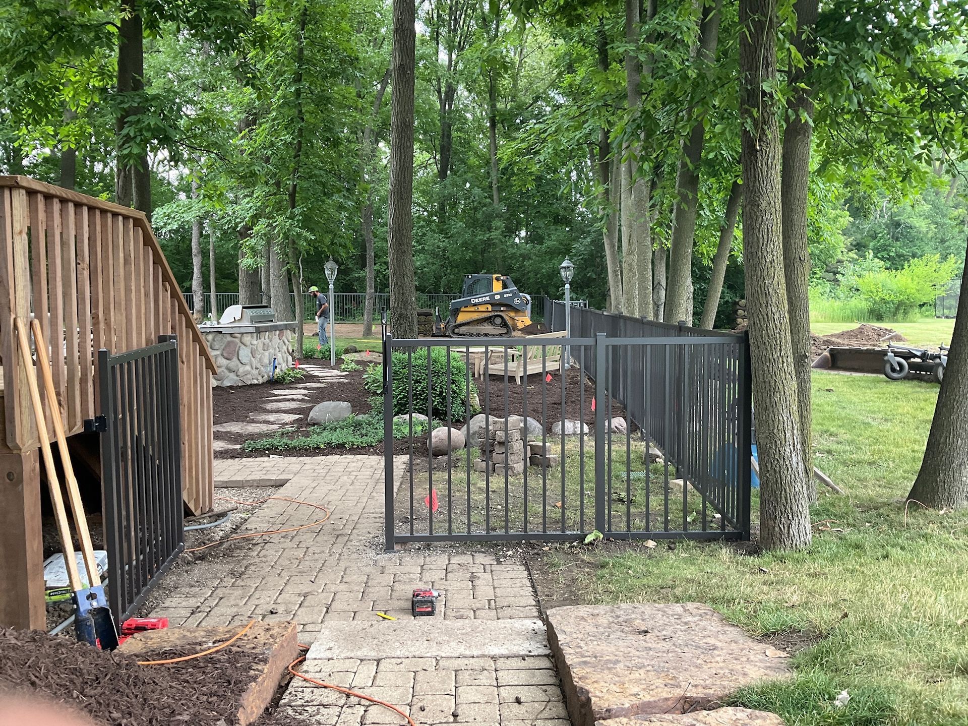 A backyard scene with a stone pathway, a metal fence, trees, and landscaping equipment.