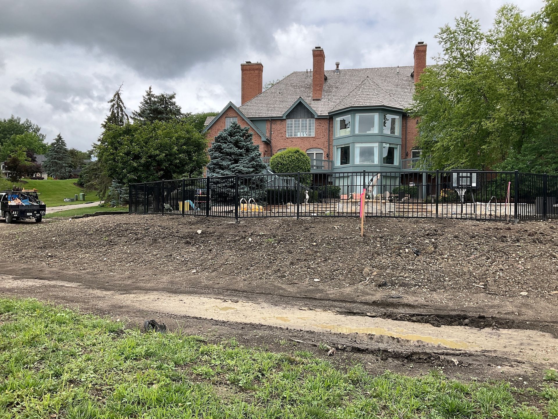 Large house behind a black fence. Dirt and gravel in the foreground. Construction vehicle on the left.