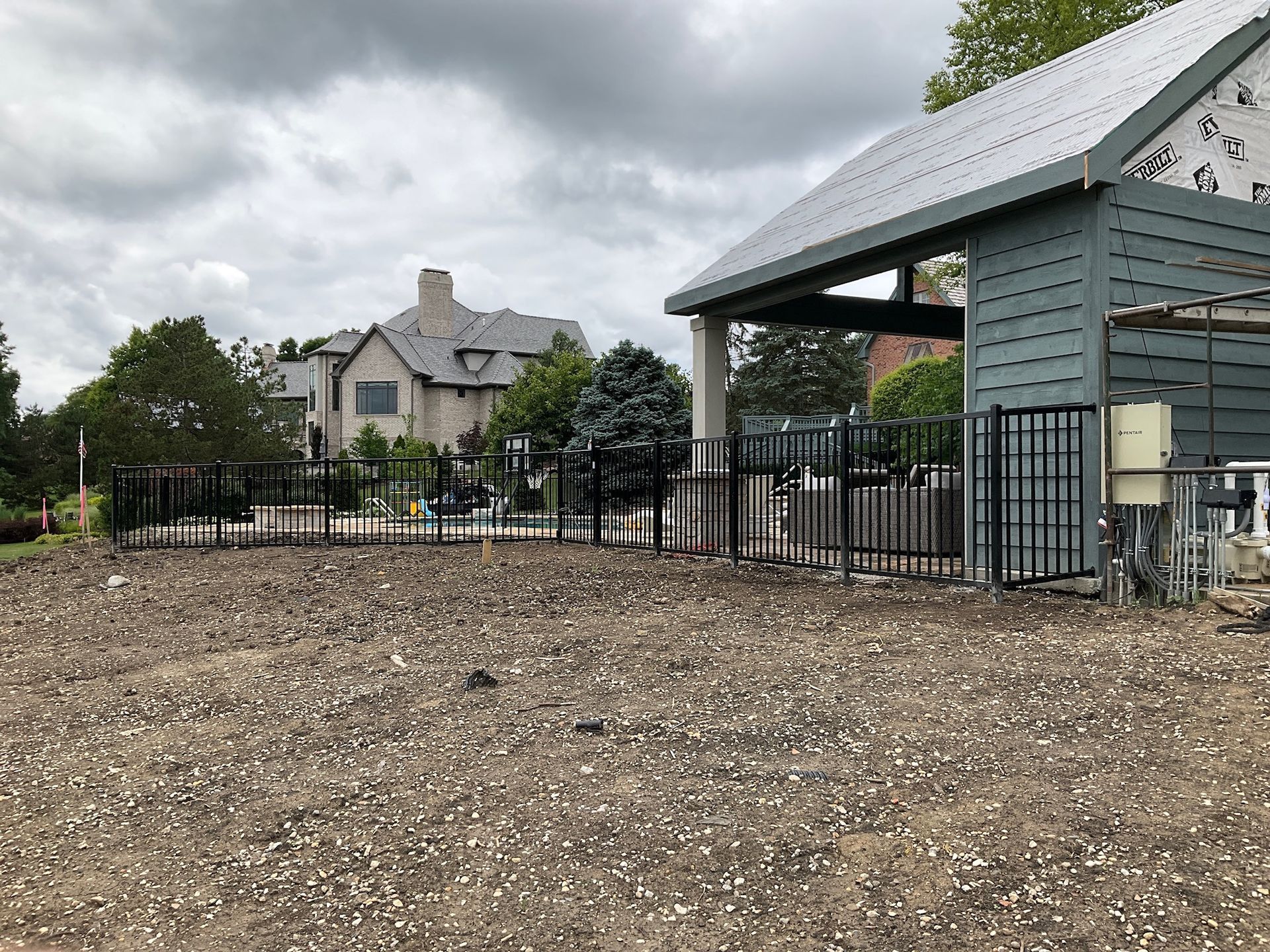 Gravel lot with a partially built shelter and black metal fence in front of a house under cloudy skies.