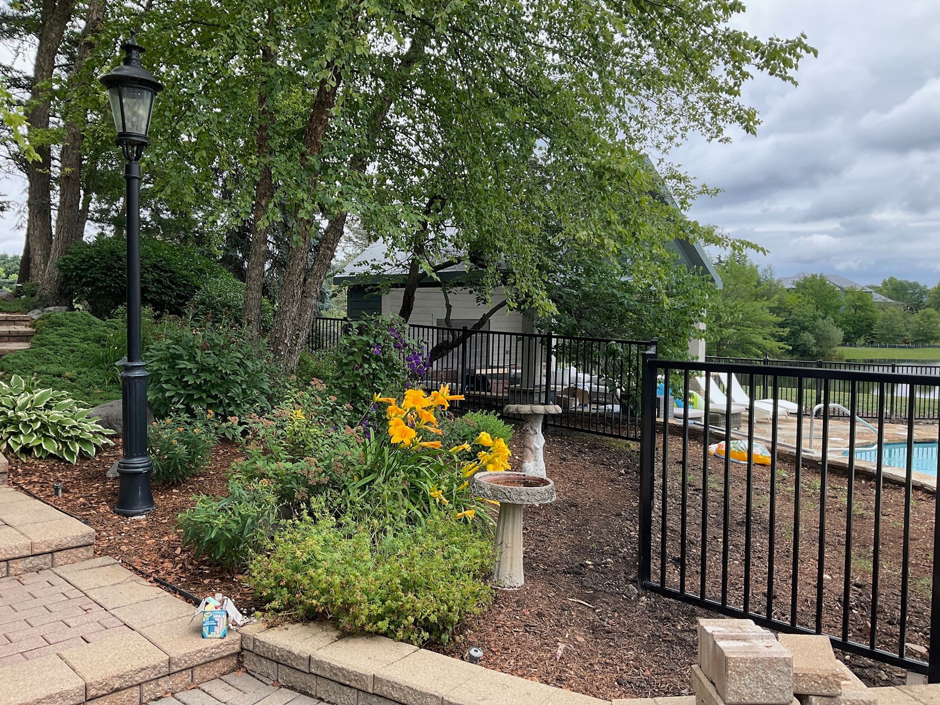 Backyard with pool, black fence, and garden with flowers. Lamp post, tree, and cloudy sky visible.