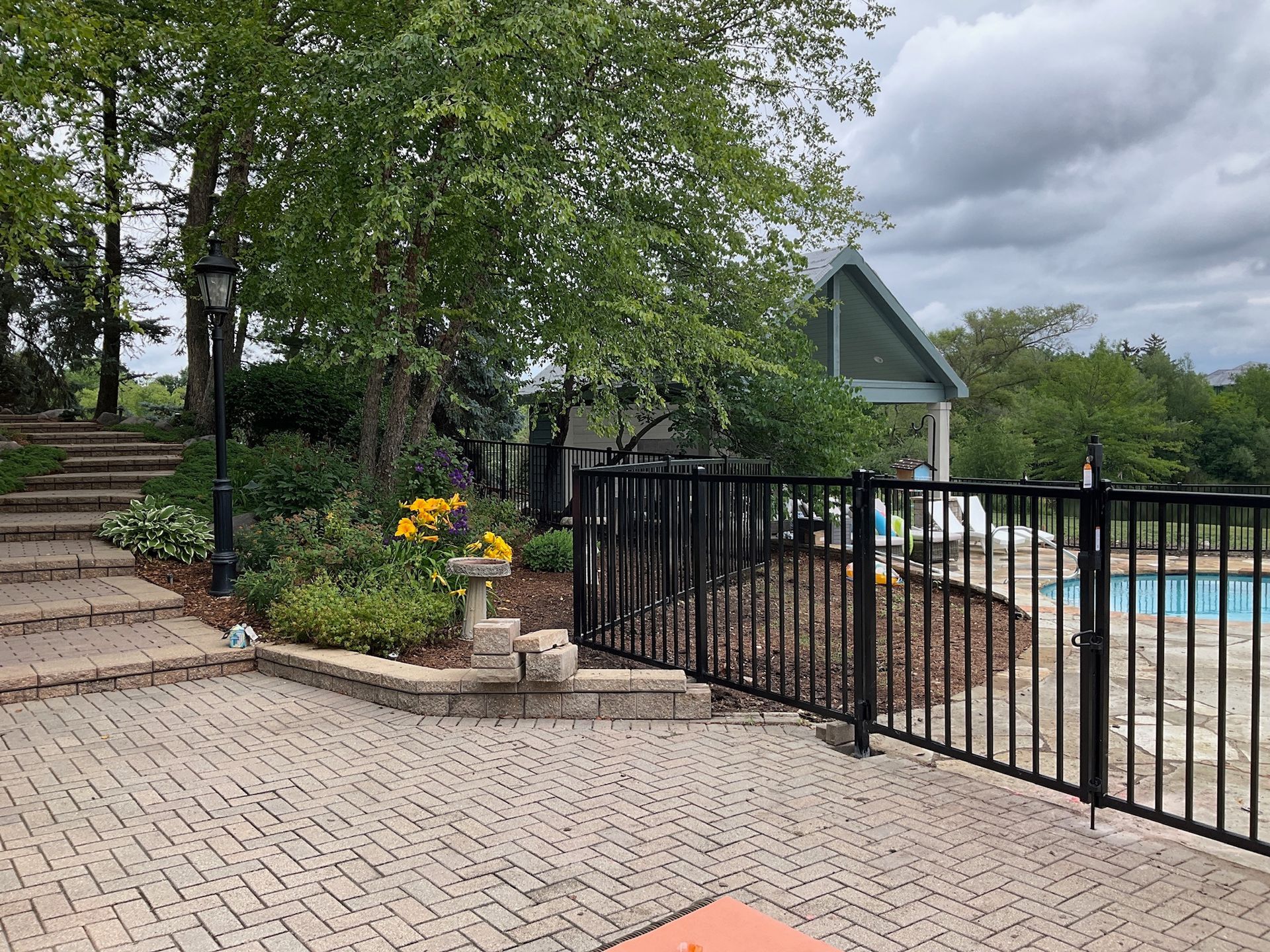 Brick patio with a black fence bordering a pool, greenery, and steps leading upwards.