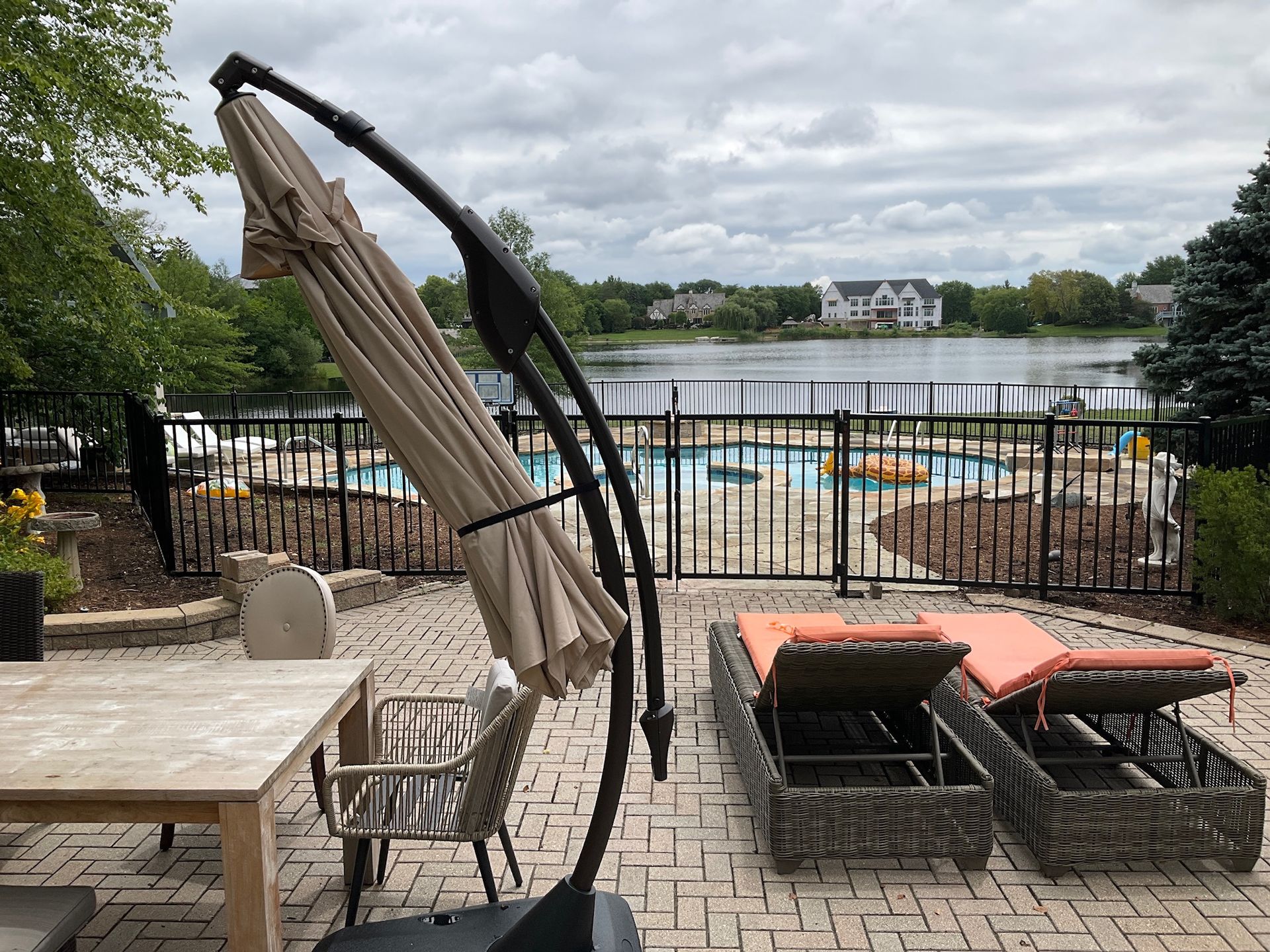 Patio with a lake view; closed beige umbrella, lounge chairs, table, and a black fence.