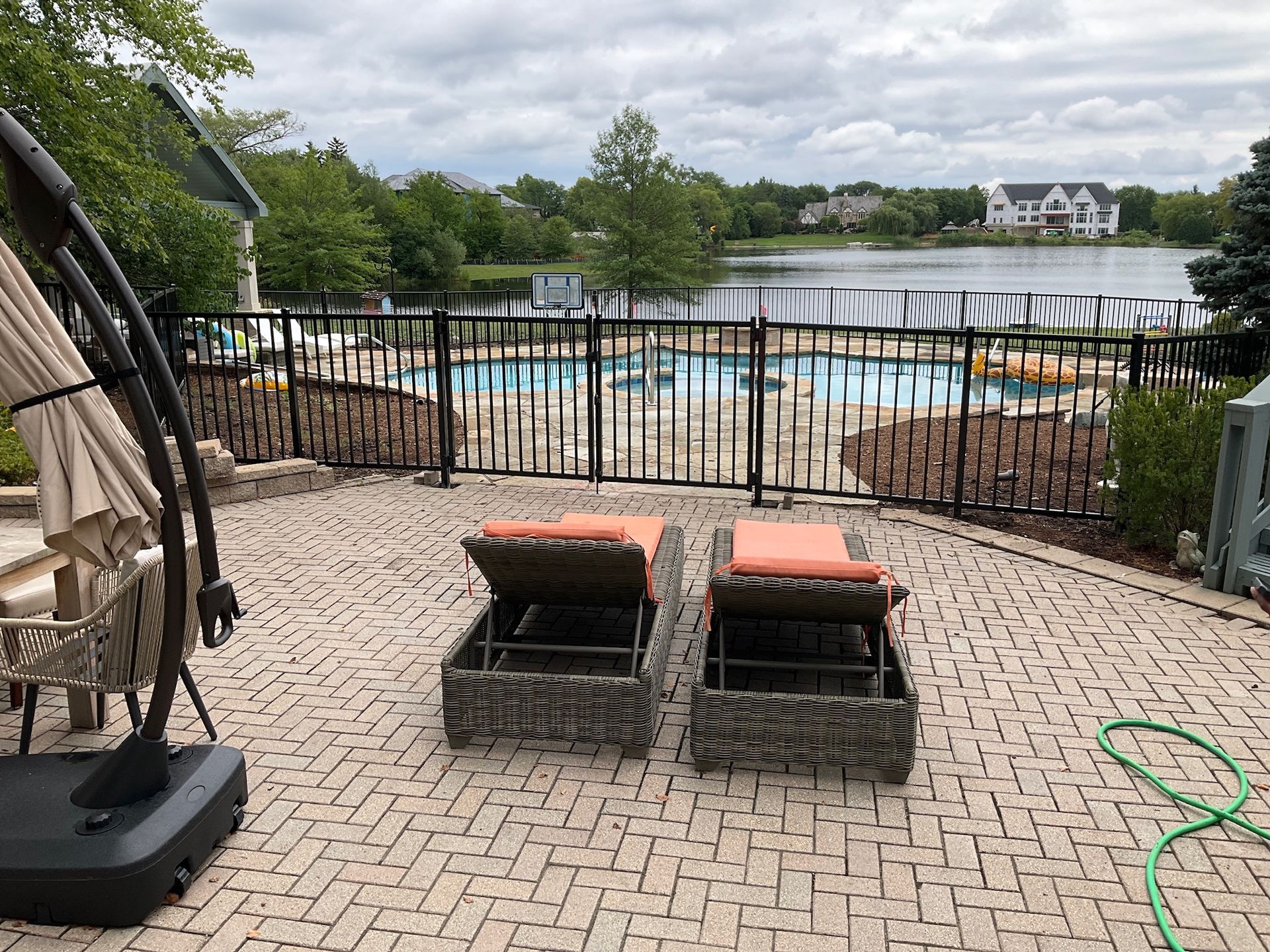 Brick patio with two lounge chairs, black fence around pool, lake in background.