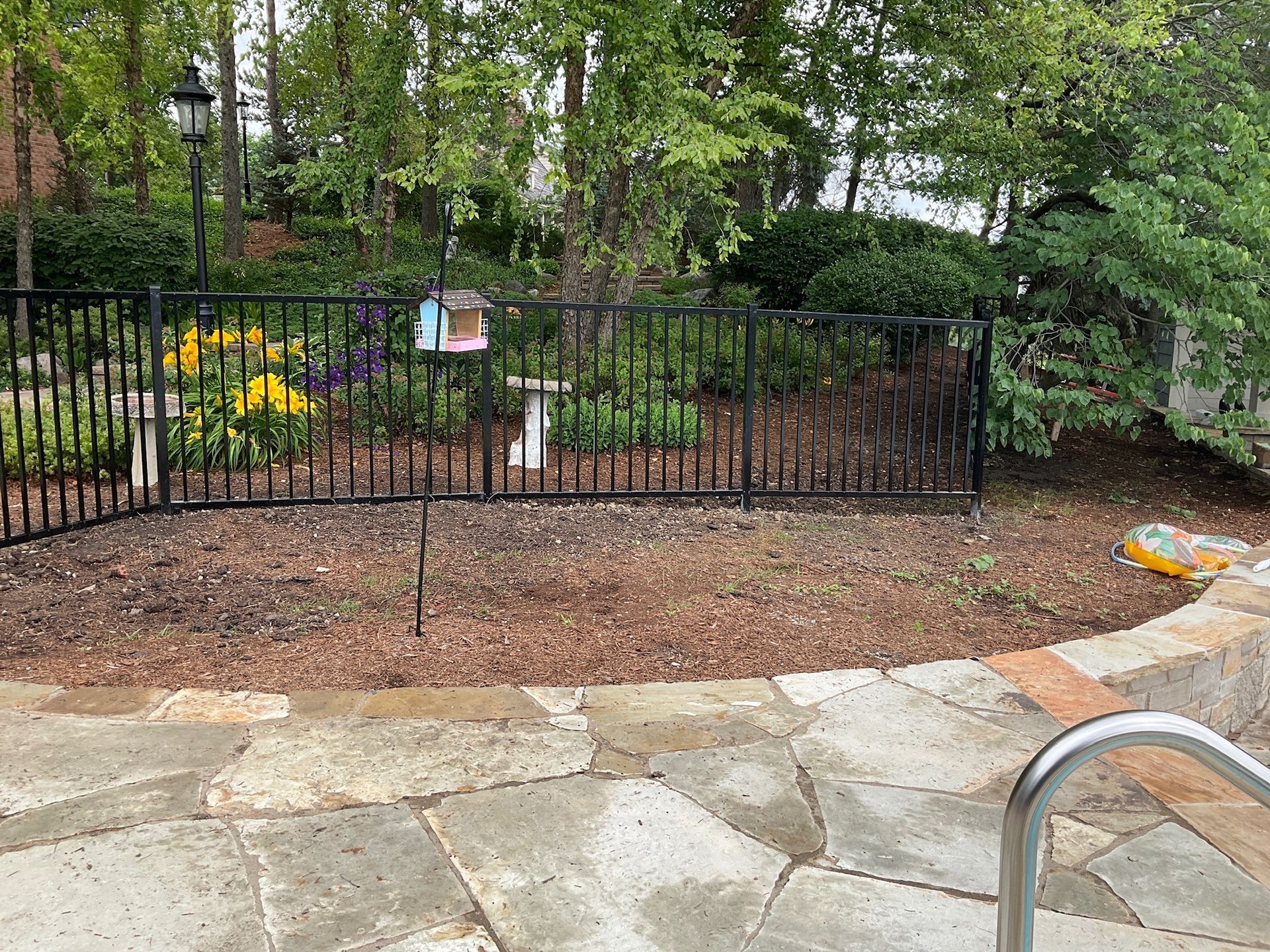 Black fence surrounds a flowerbed with a birdhouse, next to a stone patio and a pool edge.