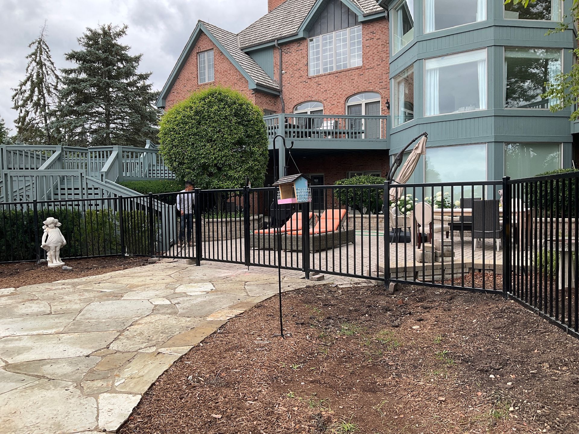 Black metal fence encloses a patio and backyard with a brick house and wooden deck in the background.