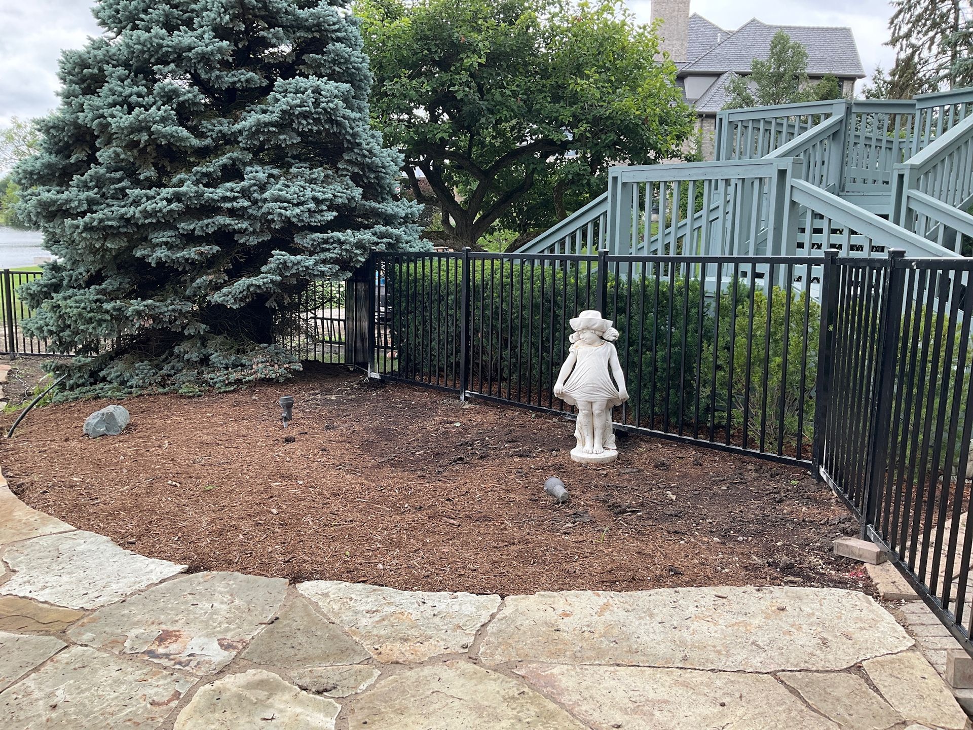Yard with a black fence, a blue spruce tree, stone patio, and a statue.