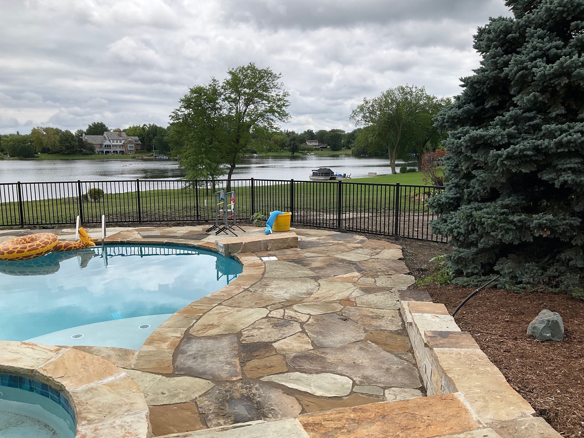 Poolside view overlooking a lake with black fence and cloudy sky. Stone patio, blue pool, and evergreens.