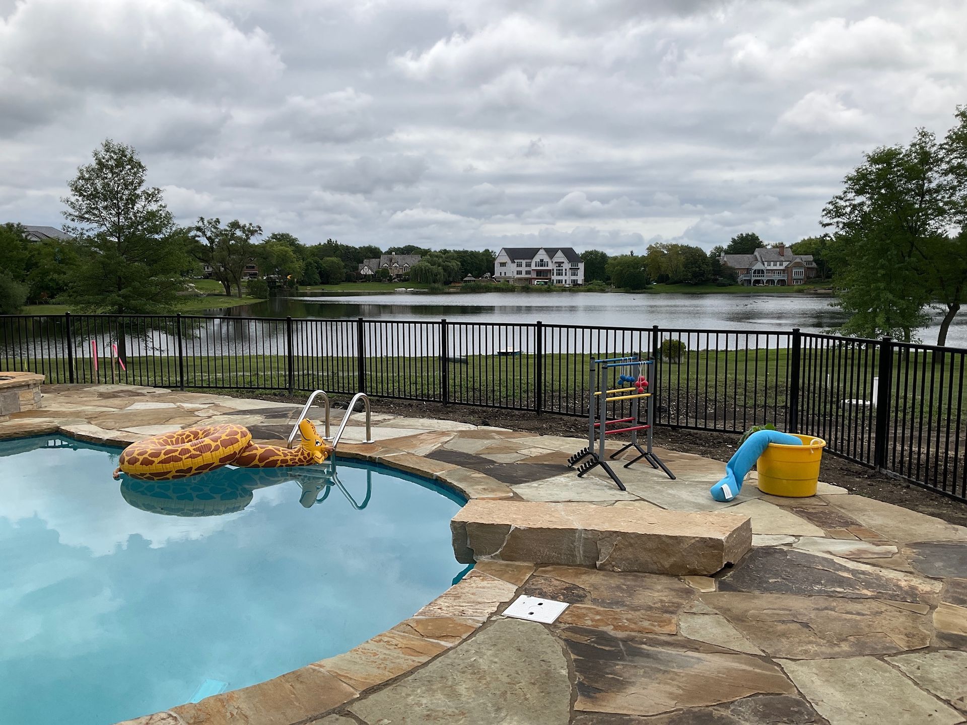 Poolside view of a lake and houses under a cloudy sky. Black fence, rocks, and equipment are visible.