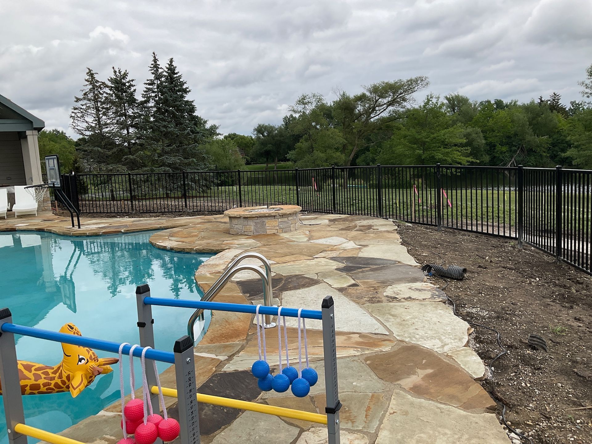 Poolside area with a pool, stone patio, fire pit, and fence under a cloudy sky.