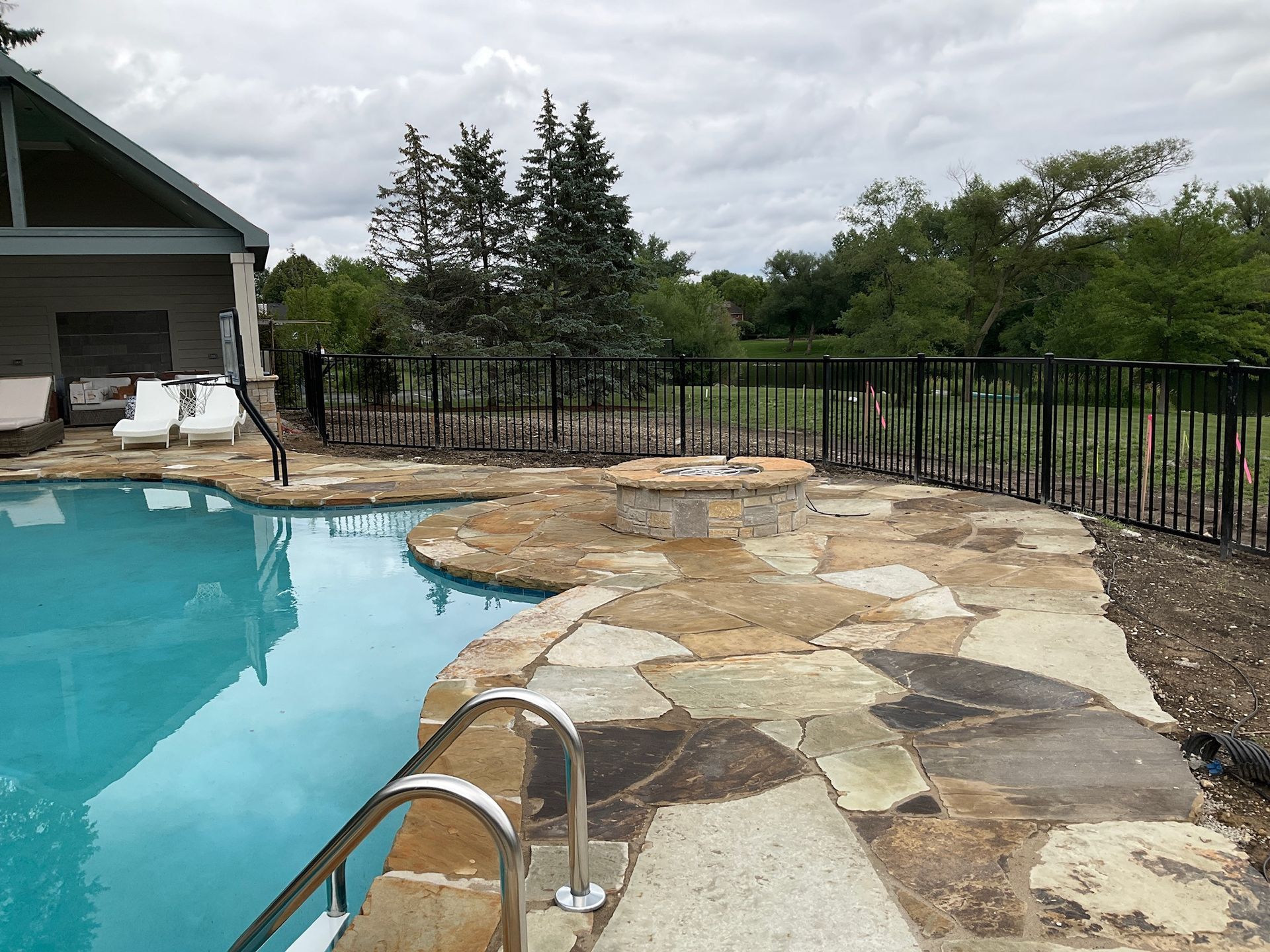 Pool with stone patio, fire pit, black fence, and trees under a cloudy sky.
