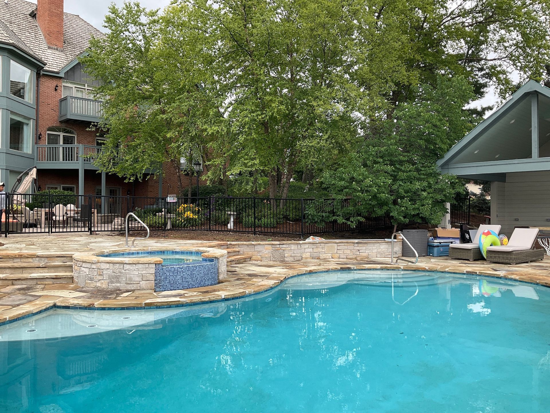 Pool and hot tub with stone surround, trees, and house in background.