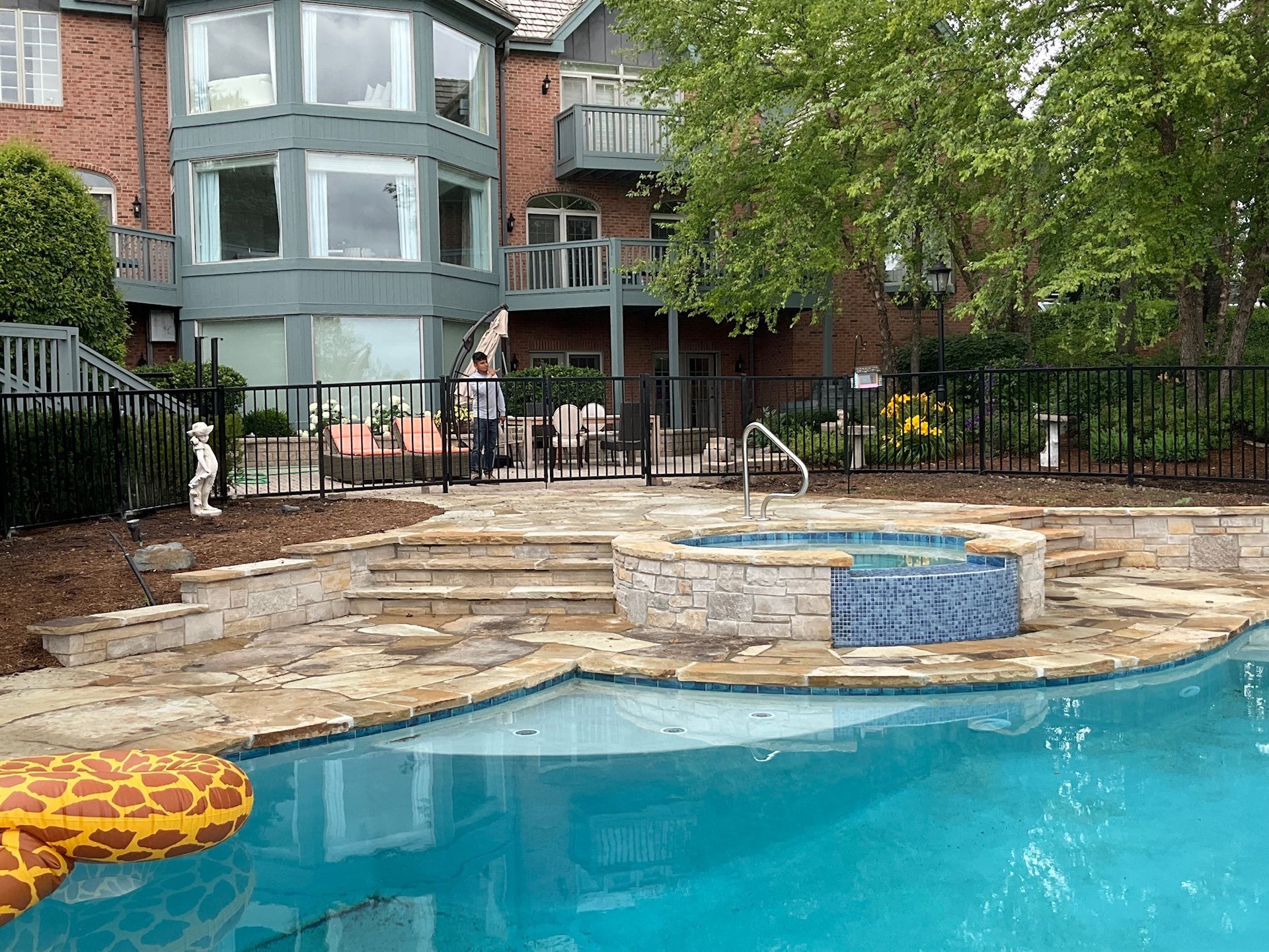 Pool area with steps, hot tub, and house in the background. Green trees and black fence.