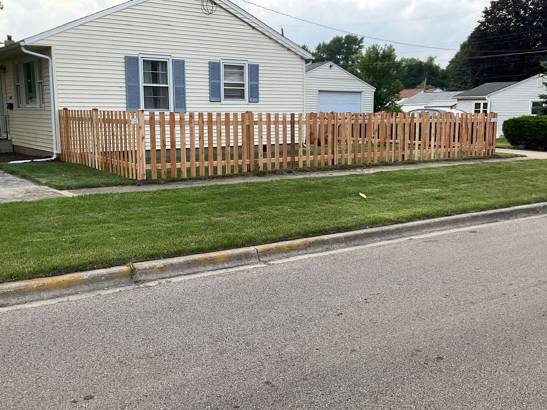 A low, wooden picket fence surrounds the front yard of a light-colored house with blue shutters.