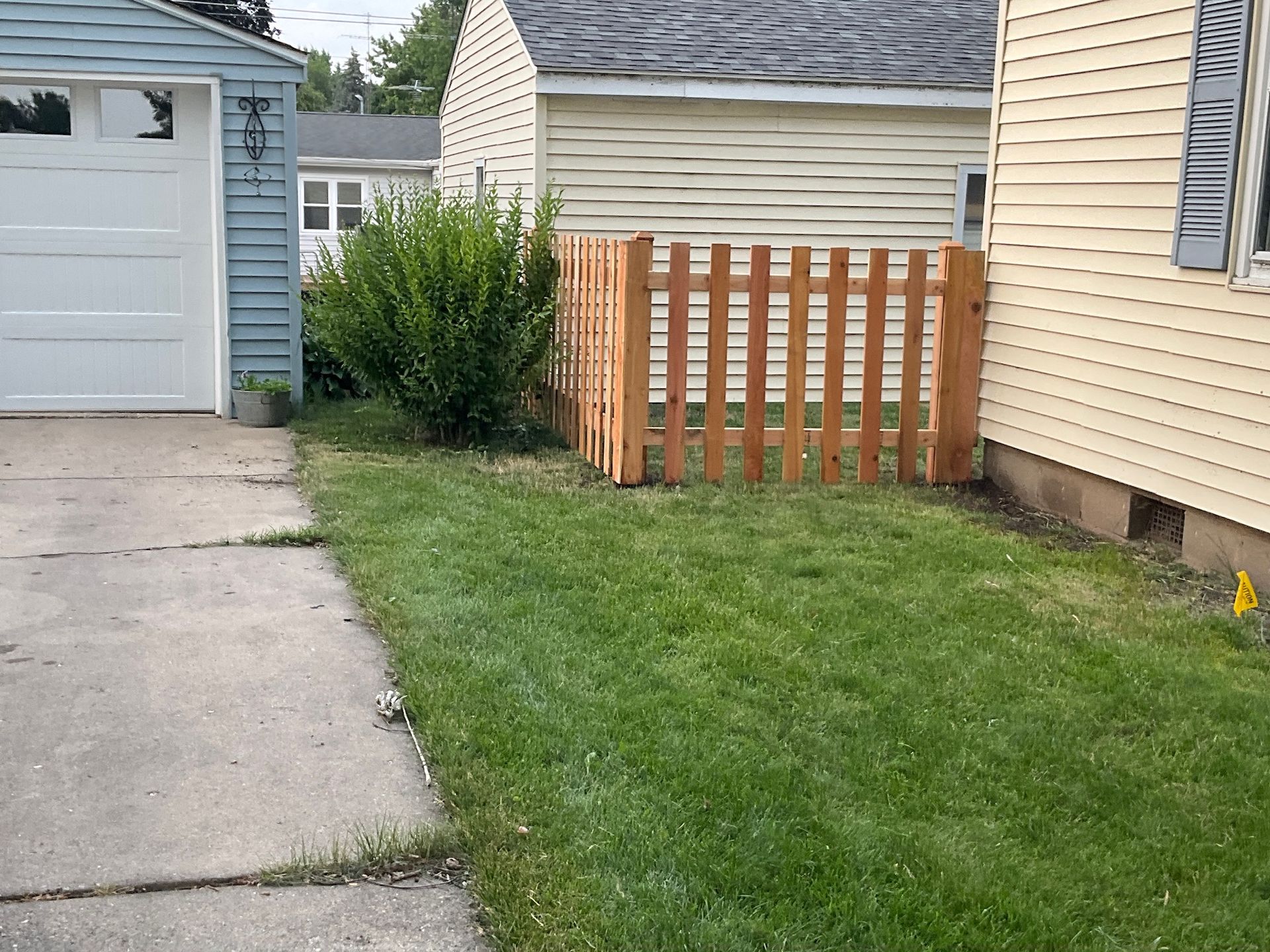 Wooden fence around a bush next to a house and garage; grass lawn.