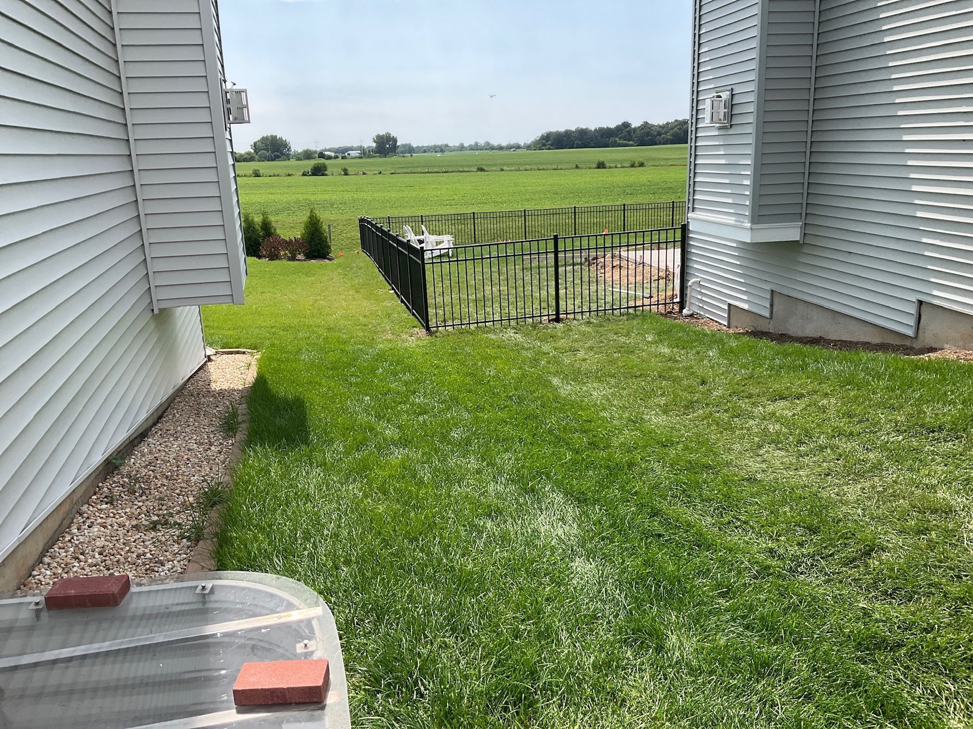 Lawn between two white buildings leads to a black fence bordering a field on a sunny day.