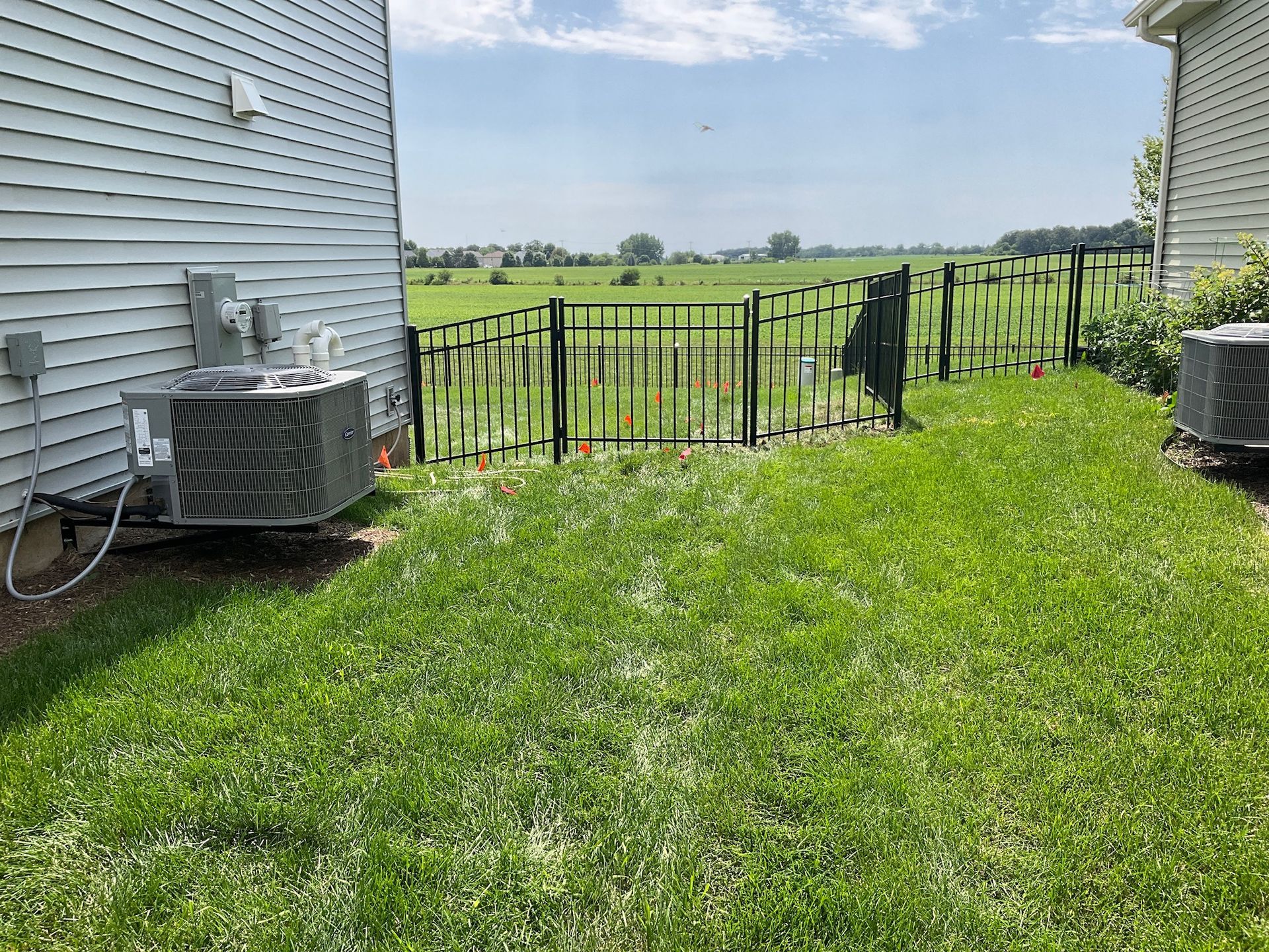 Lawn between two houses with black fence, a gate, and HVAC units. Field in background under a sunny sky.