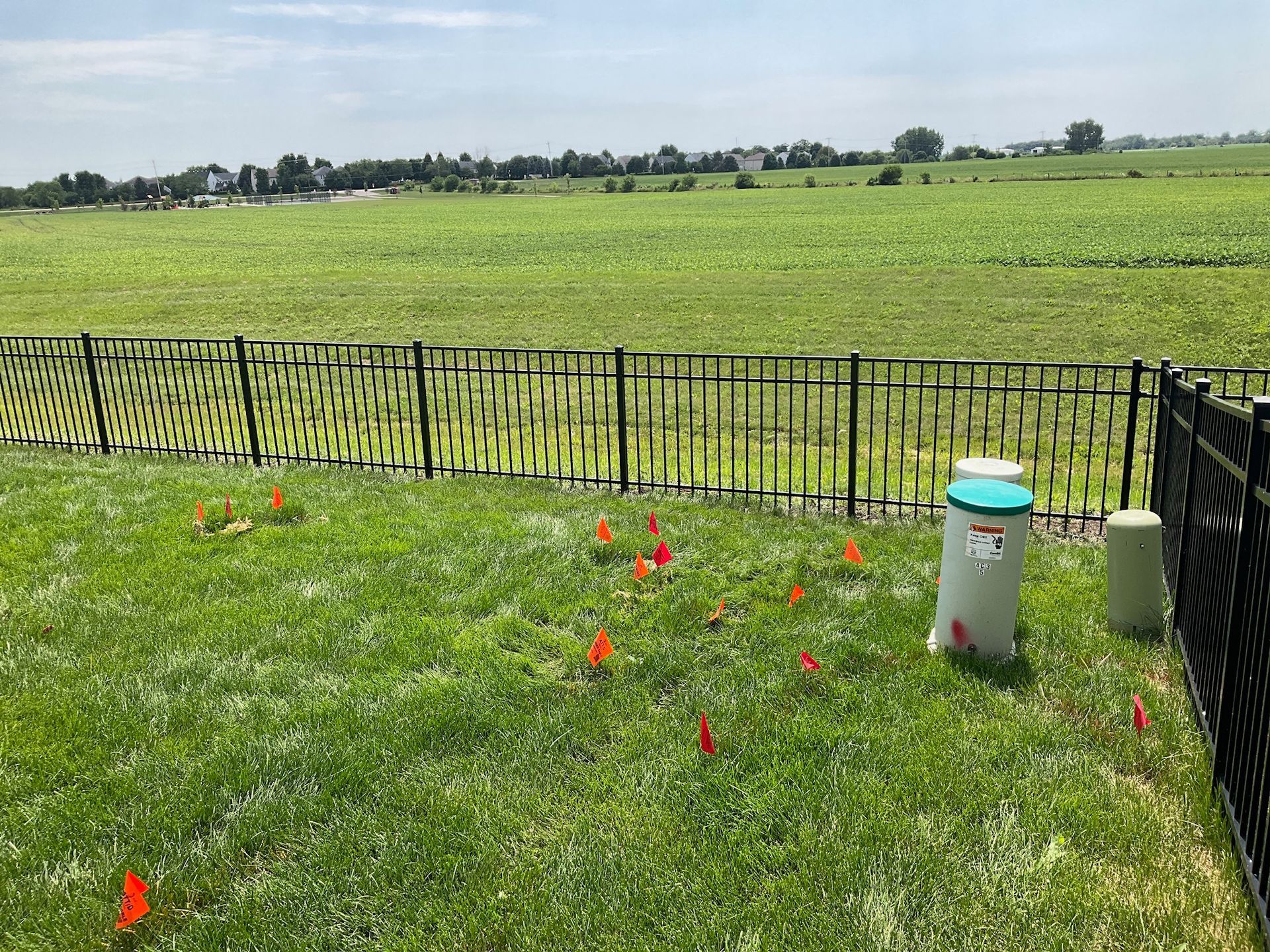 Green lawn with orange flags, black fence, and utility access points, with a field in the background.