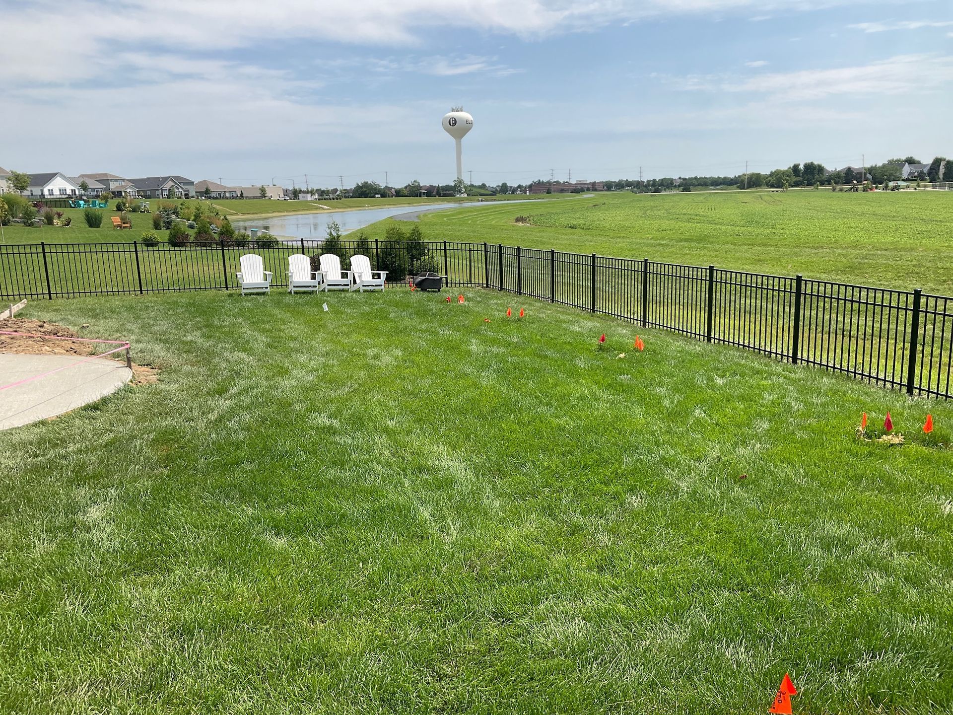 Green lawn with black fence, chairs, and a water tower in the background under a cloudy sky.